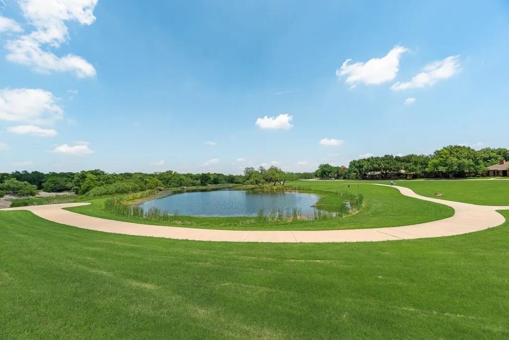 Expansive green space featuring a tranquil pond, a winding paved pathway, and mature trees creating a natural border