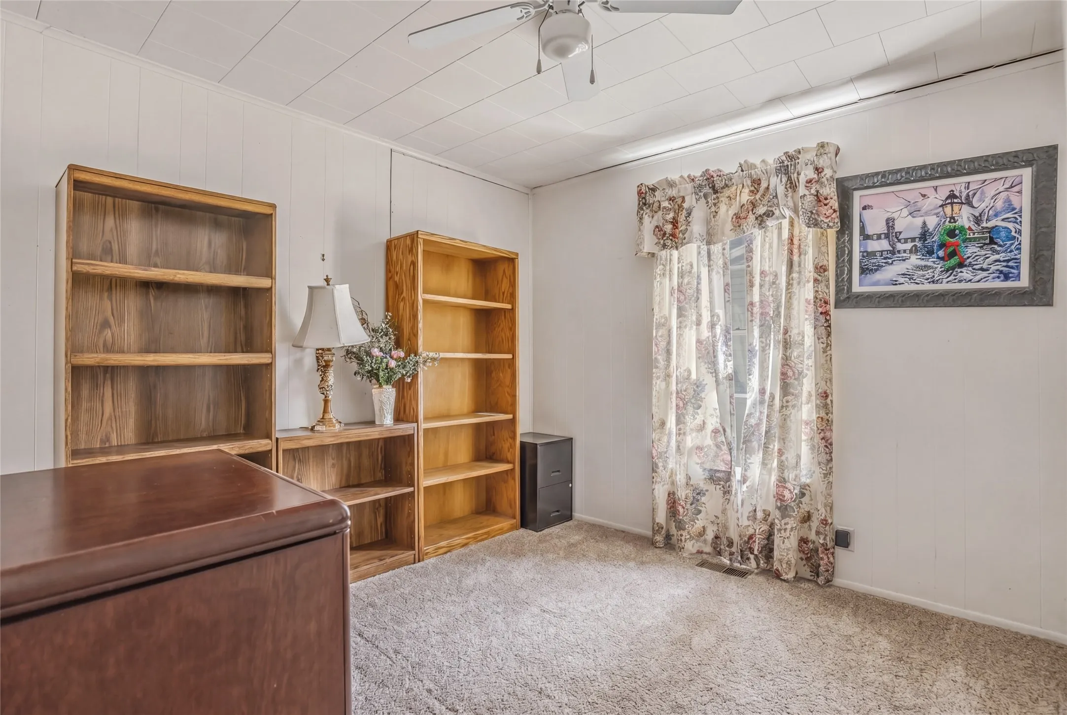 Carpeted room featuring white paneling, a drop ceiling, and a window with floral drapes