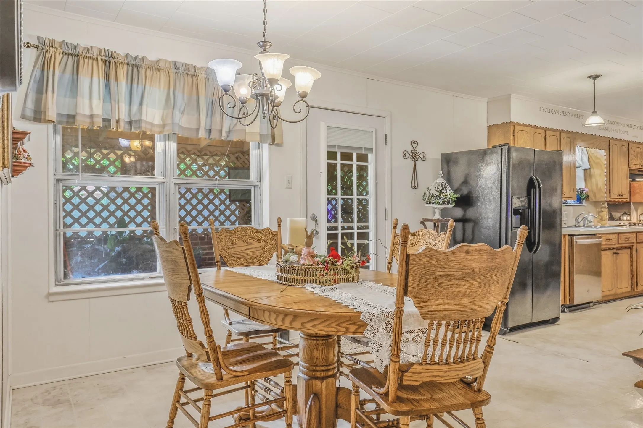 Dining area featuring an overhead chandelier, two windows with lattice-patterned screens, and an adjacent kitchen with wood cabinetry and stainless steel appliances