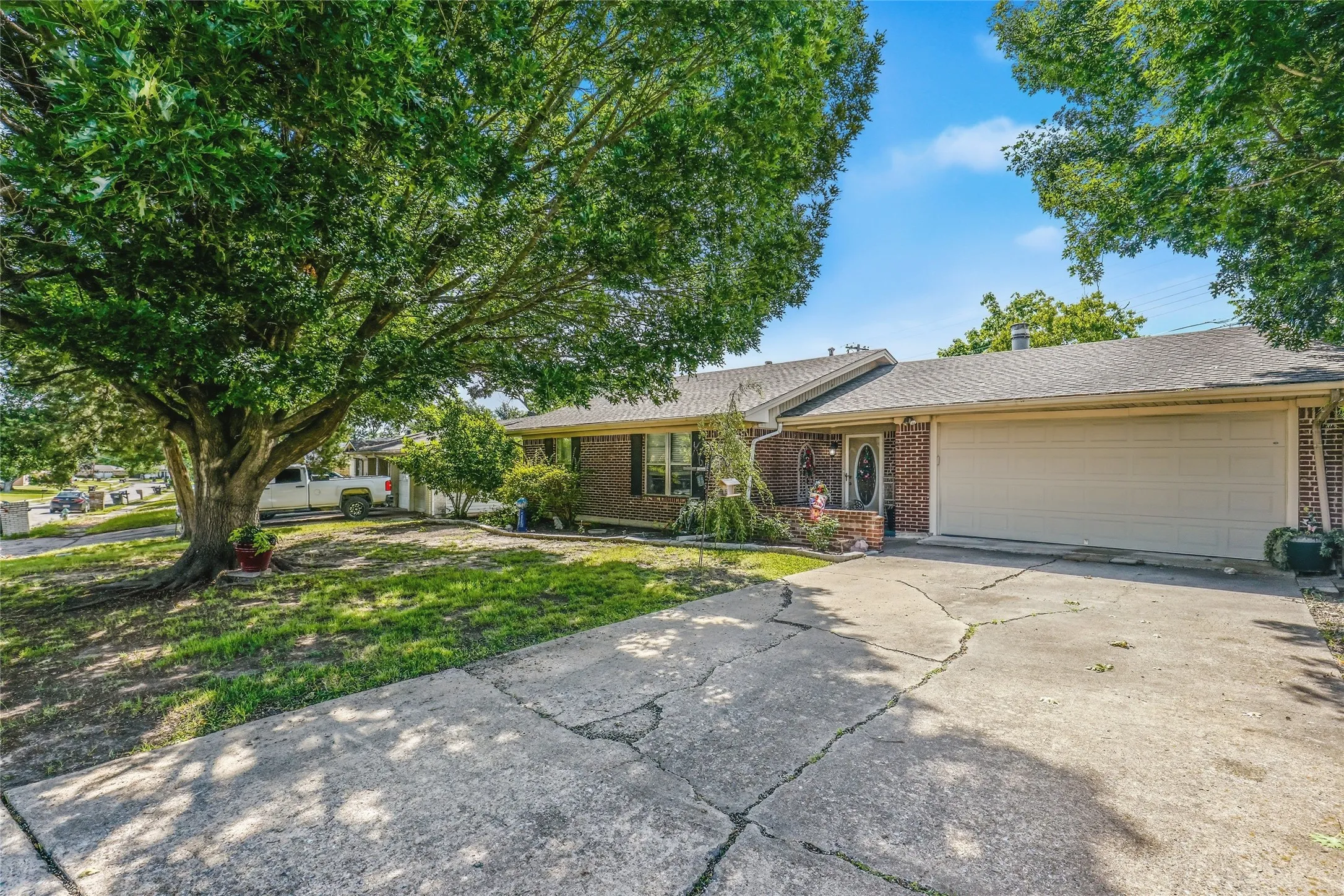 Brick-front single-story residence featuring a two-car garage, concrete driveway, and established front yard landscaping with mature trees
