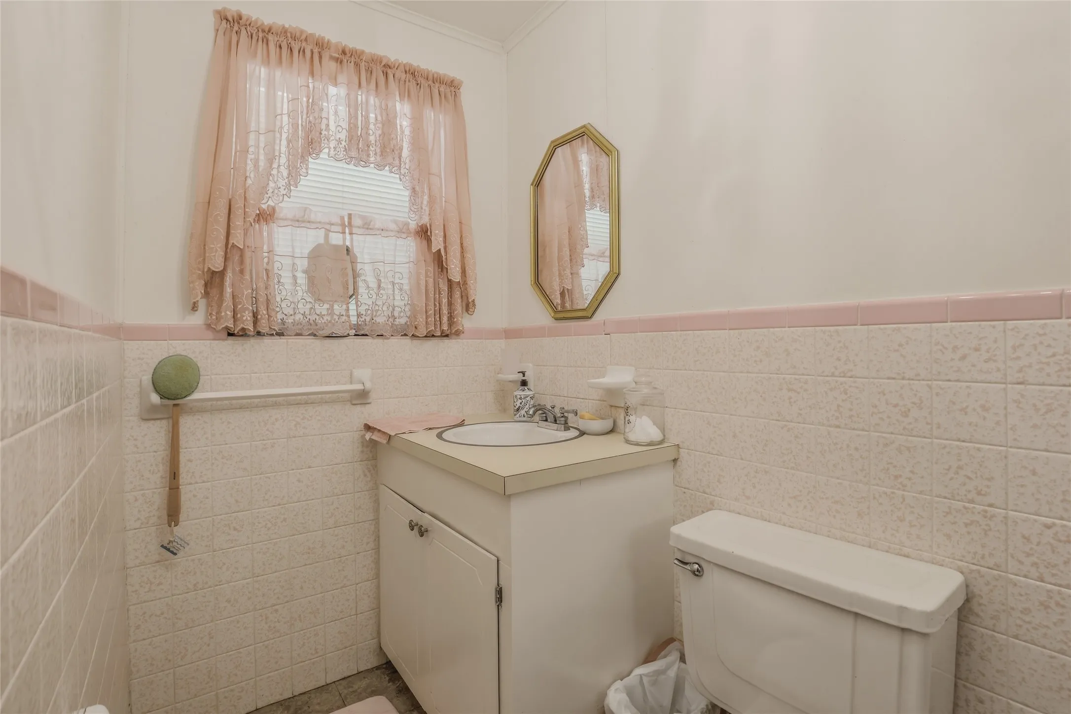 Bathroom featuring a vanity with an integrated sink, a toilet, an octagonal mirror with a gold-tone frame, and tiled walls with a pink accent stripe