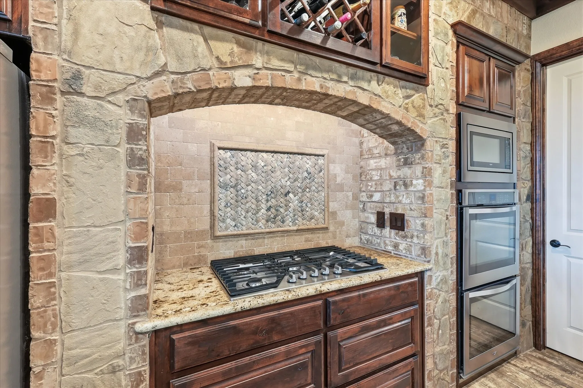 Stone and brick kitchen alcove featuring a gas cooktop, granite countertop, rich wood cabinetry, and stainless steel built-in ovens
