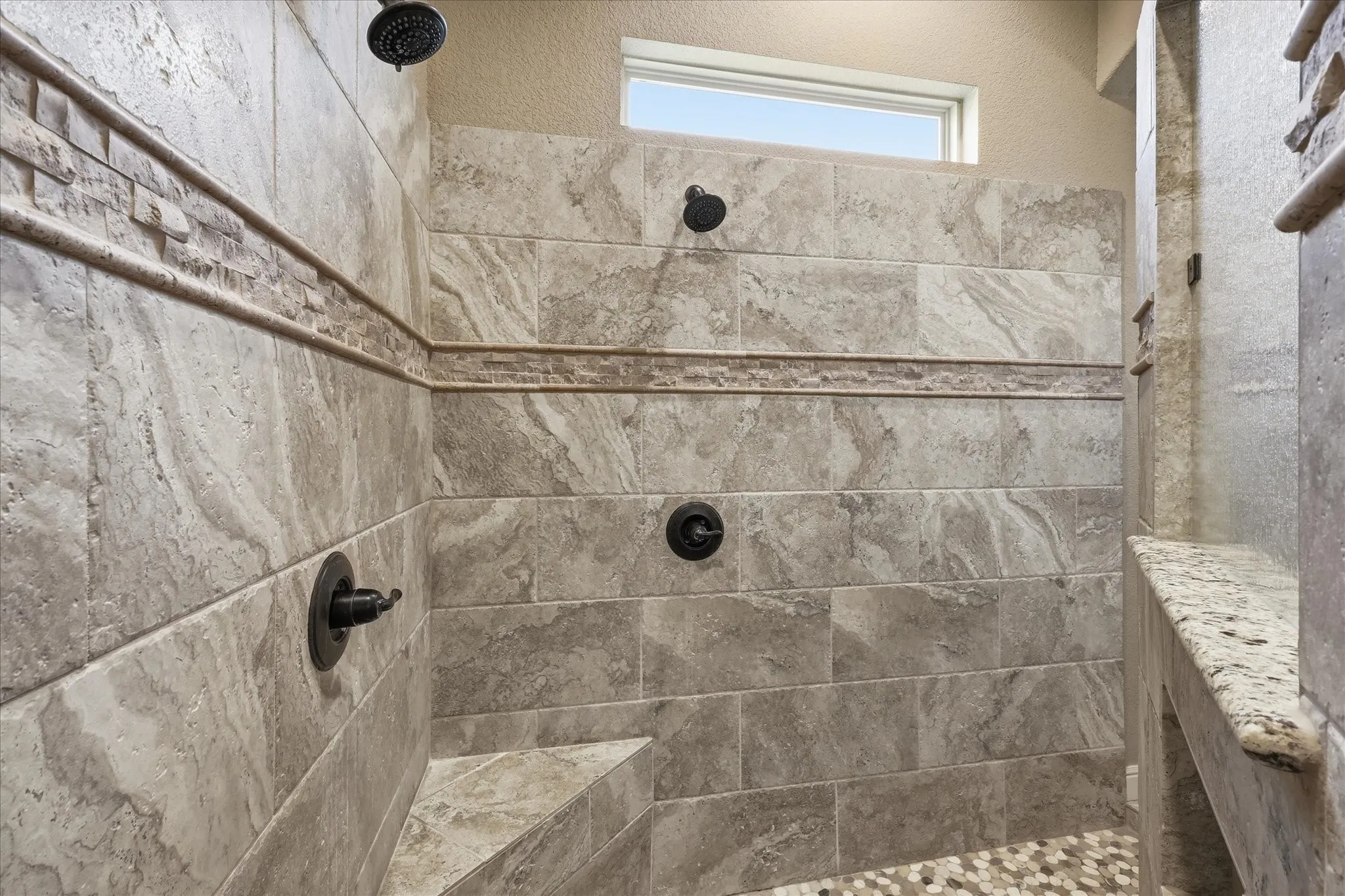 Tiled shower enclosure featuring dual shower heads with dark bronze fixtures, an integrated tiled bench, and a window for natural light