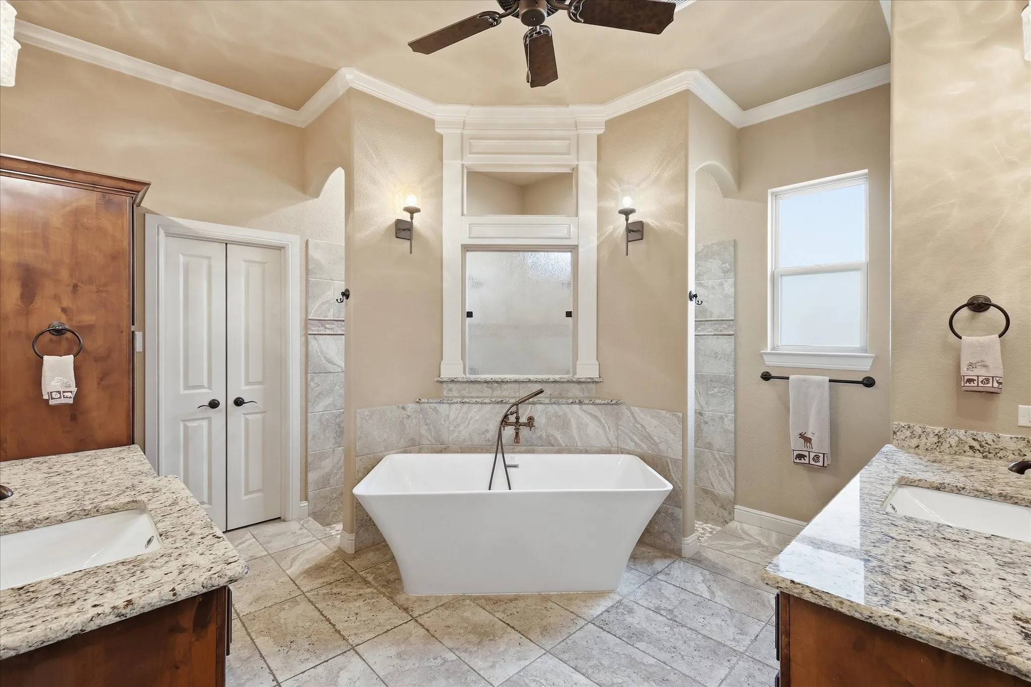 Freestanding soaking tub with a floor-mounted faucet, dual vanities with granite countertops, tiled flooring, and a walk-in shower with a glass enclosure