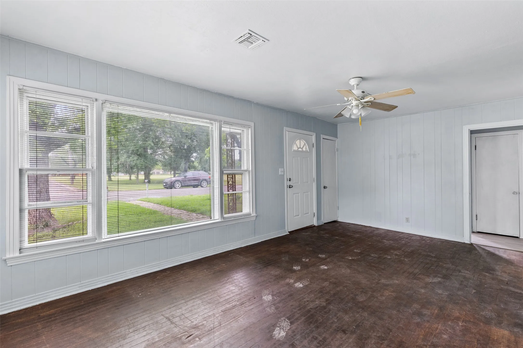 Living area featuring a large picture window, two side windows with blinds, wood-finish flooring, a ceiling fan with light fixture, and a white paneled entry door