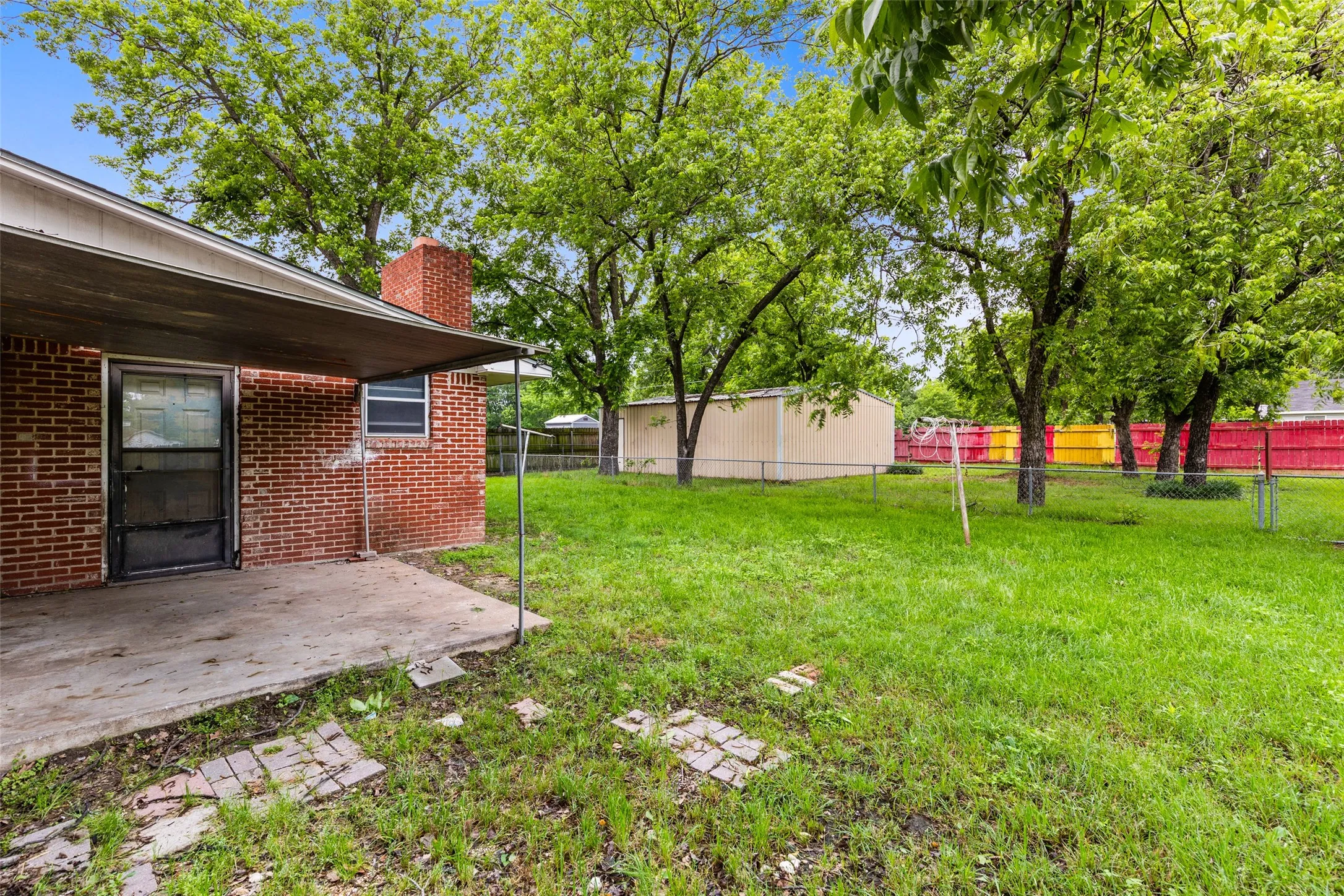 Expansive grassy yard featuring mature trees, a detached storage shed, and a covered concrete patio