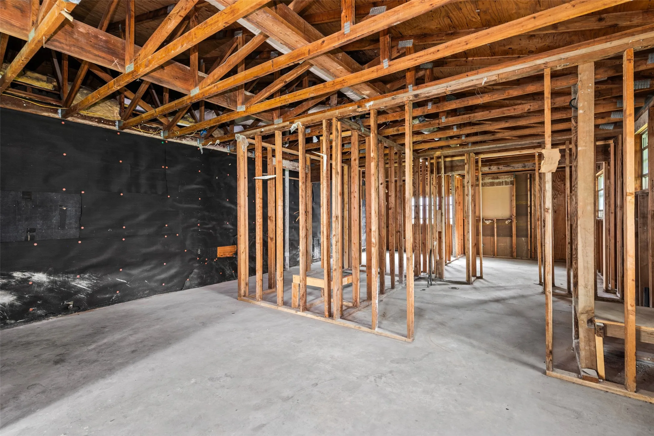 Exposed ceiling rafters and wall studs within a framed interior, featuring a concrete floor and partially covered exterior wall with black sheathing