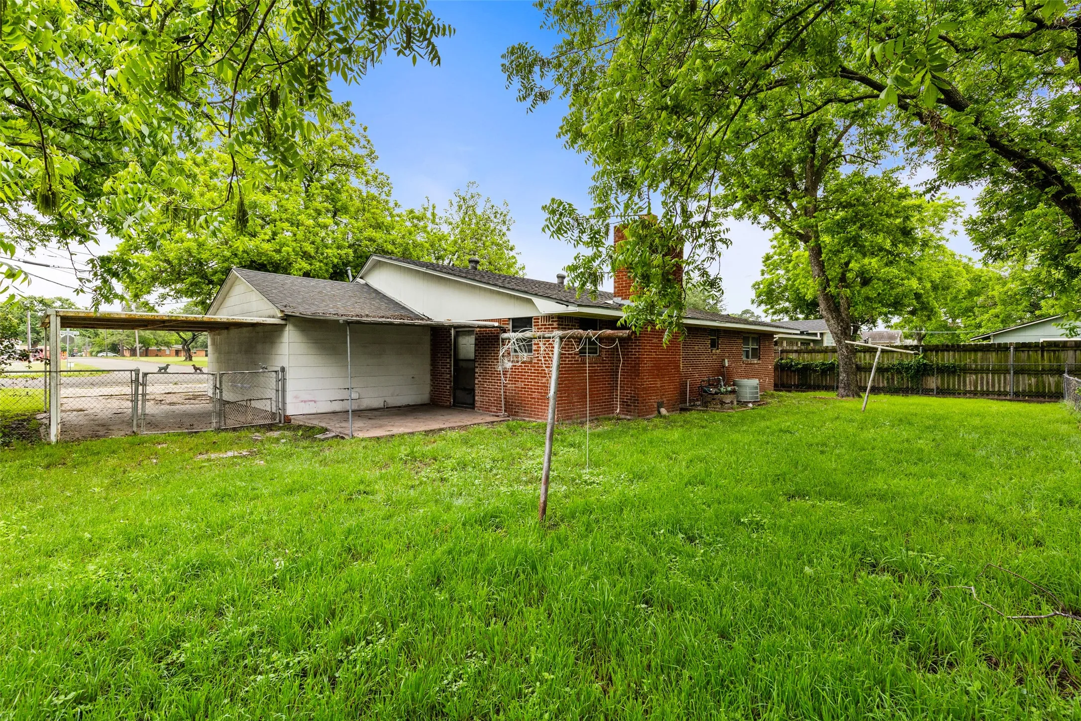 Expansive grassy backyard featuring a paved patio area, an attached carport, a brick exterior with a visible chimney, and mature trees providing natural shading