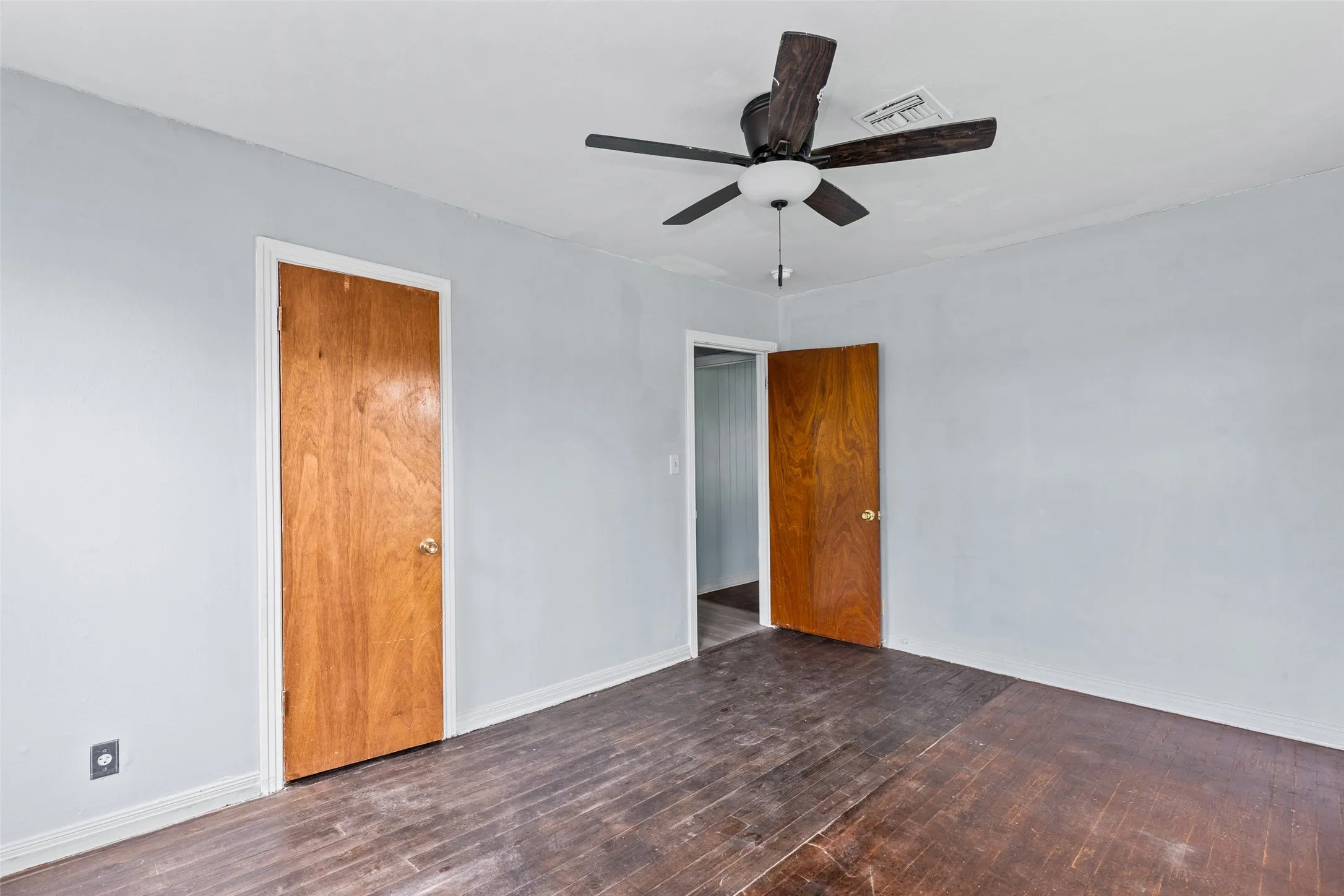 Light blue painted room featuring a ceiling fan with dark wood-finish blades, two wood-finish doors with brass-tone hardware, and dark wood-finish flooring