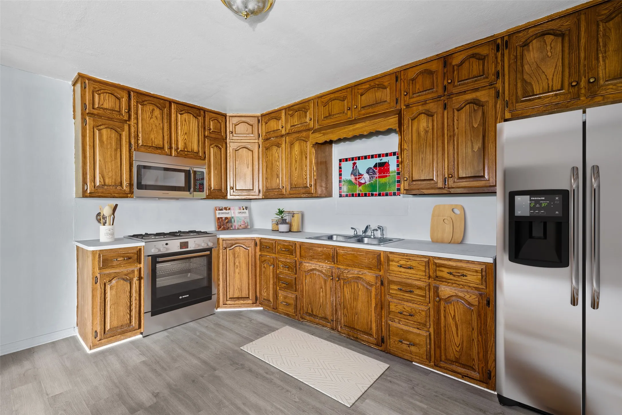 Virtual staging - Kitchen featuring wood cabinetry, light countertops, wood-finish flooring, stainless steel appliances, and an integrated stainless steel sink