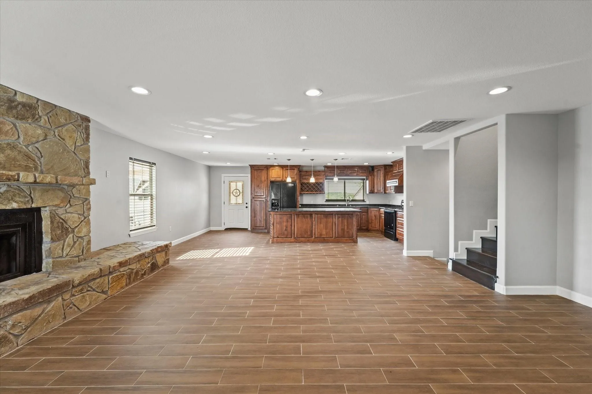 Spacious open-concept living area featuring a stone fireplace with hearth, wood-finish tile flooring, recessed lighting, and a modern kitchen with dark cabinetry and stainless steel appliances