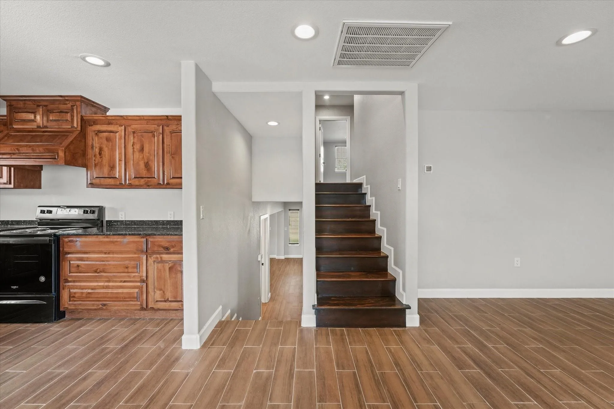 Wood-finish flooring extends throughout this space featuring a kitchen with wood cabinetry and black appliances, a staircase with rich wood treads, and recessed lighting