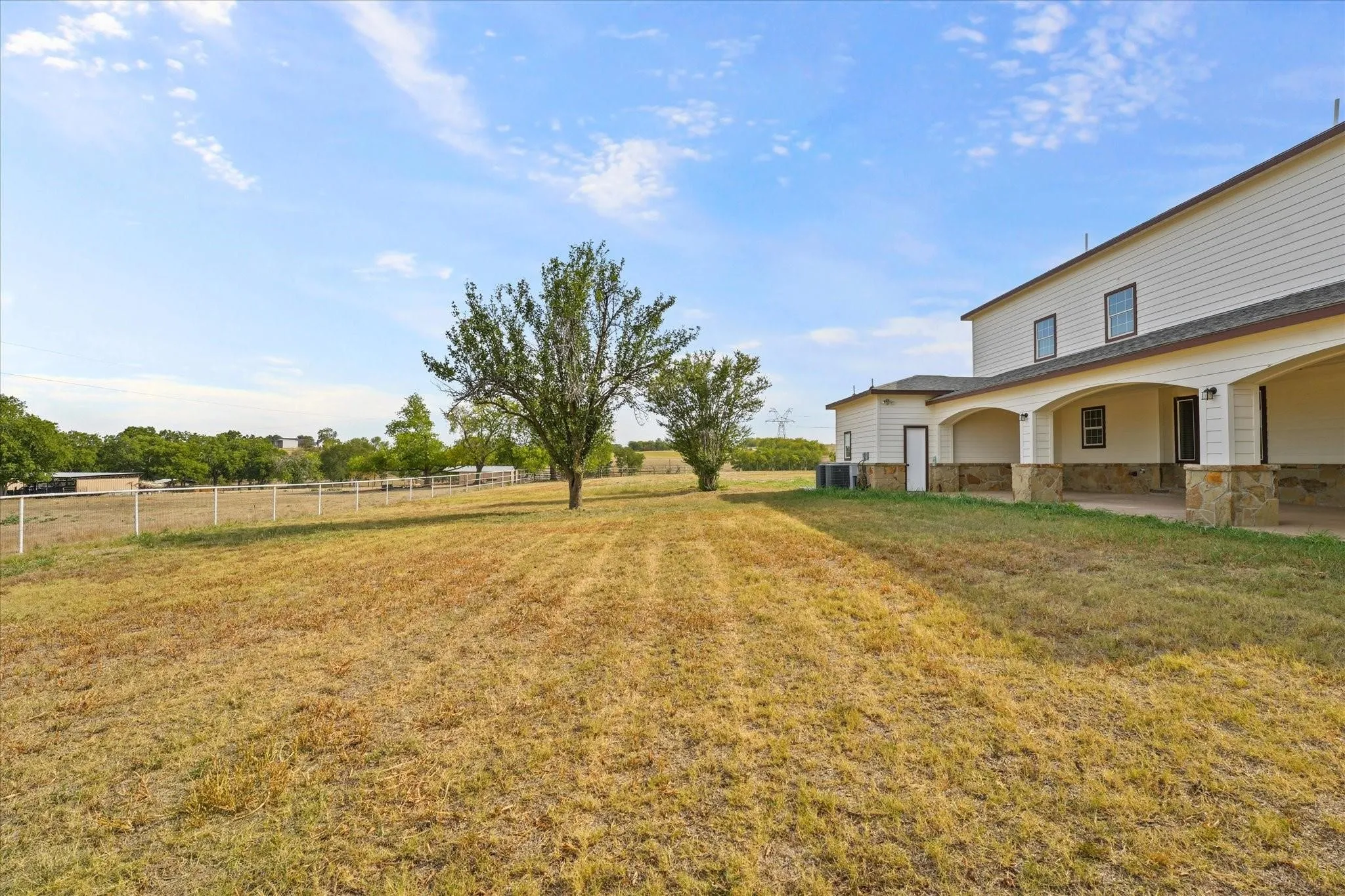 Expansive outdoor space featuring a white picket fence perimeter, mature trees, and a spacious covered patio with stone pillar accents