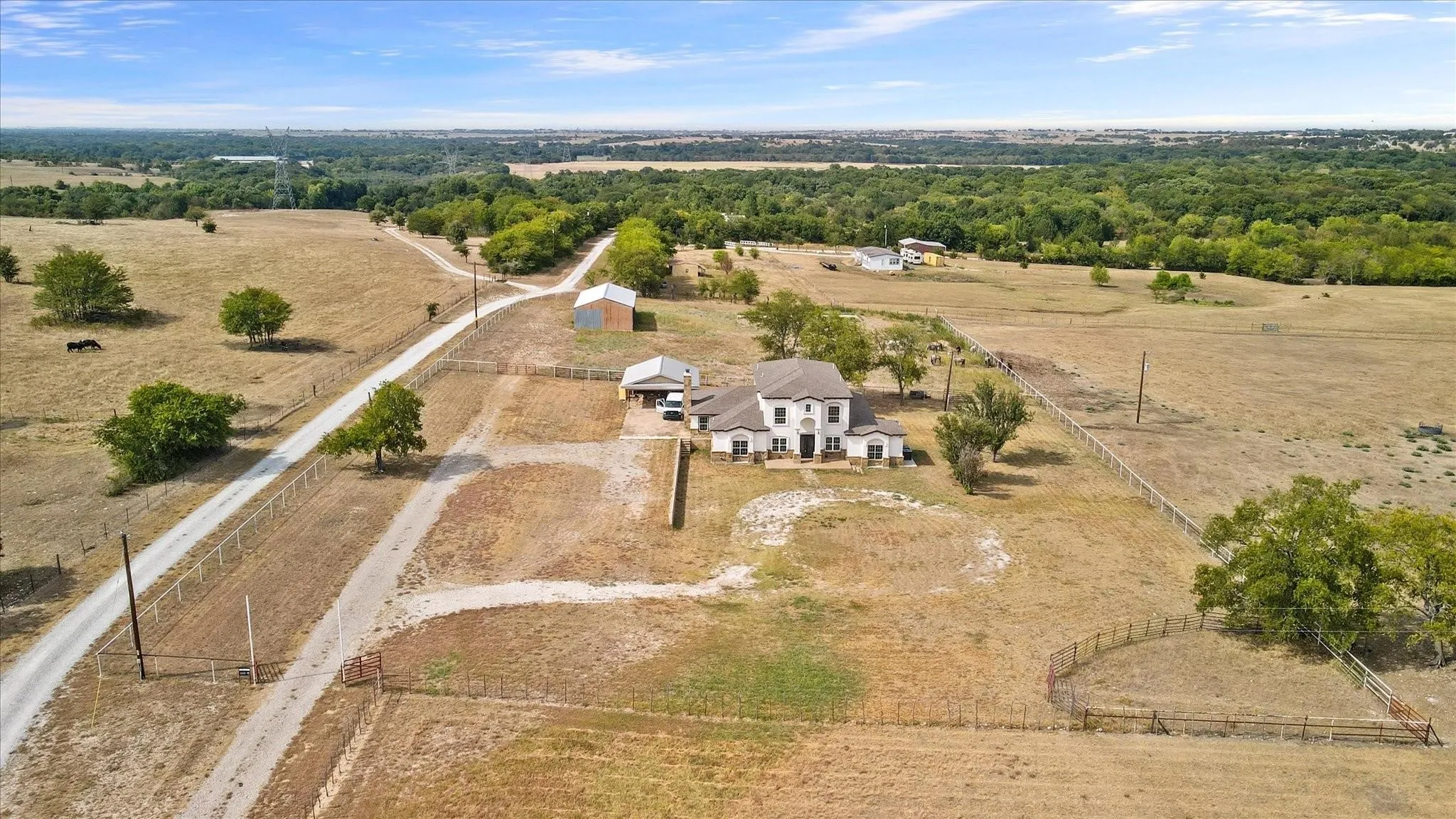 Expansive rural property featuring a main residence with a light-colored exterior and dark roof, a detached barn structure, a gravel driveway, mature trees, and fencing