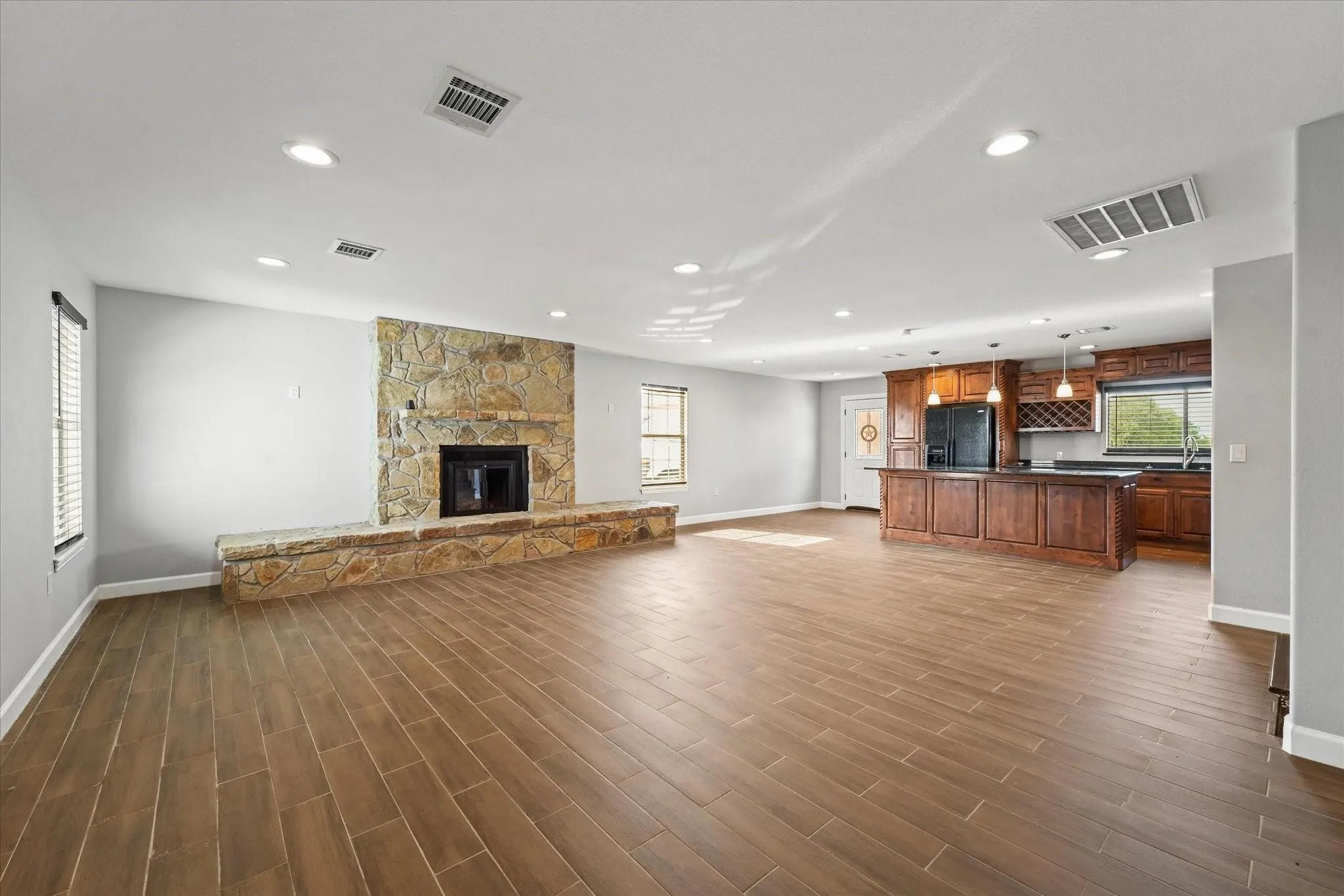 Spacious open-concept living area featuring wood-finish flooring throughout, a stone fireplace with a raised hearth, recessed lighting, and a kitchen island with pendant lighting