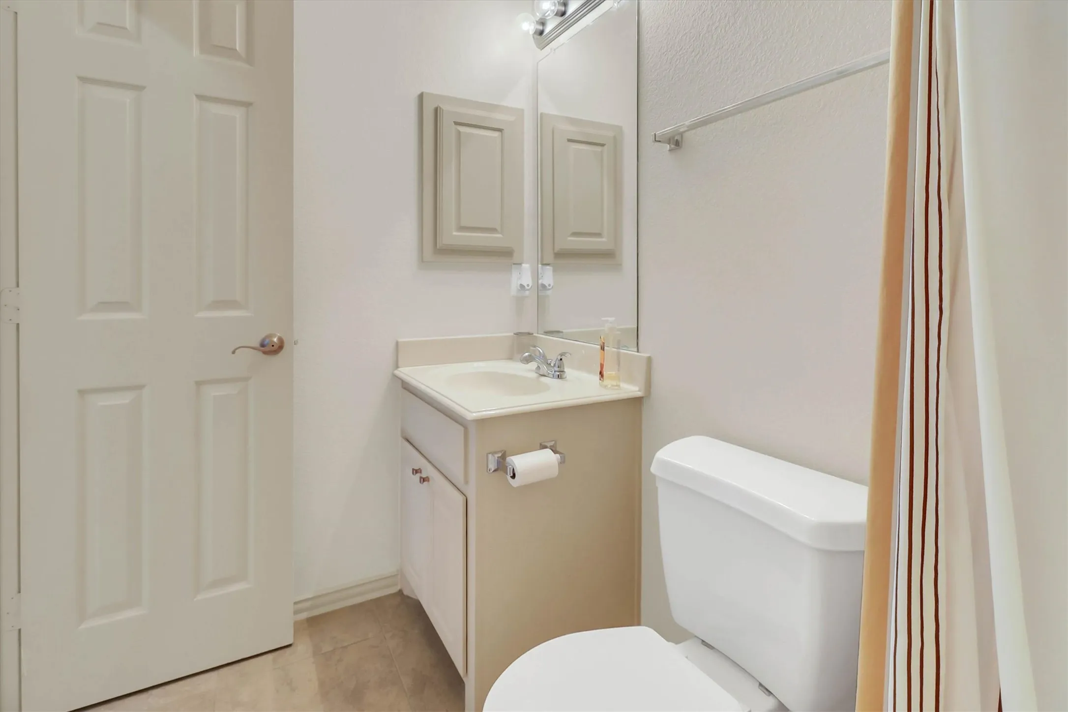 Bathroom featuring a light-colored vanity with an integrated sink, a wall-mounted mirror, overhead vanity lighting, a commode, and an adjacent paneled door