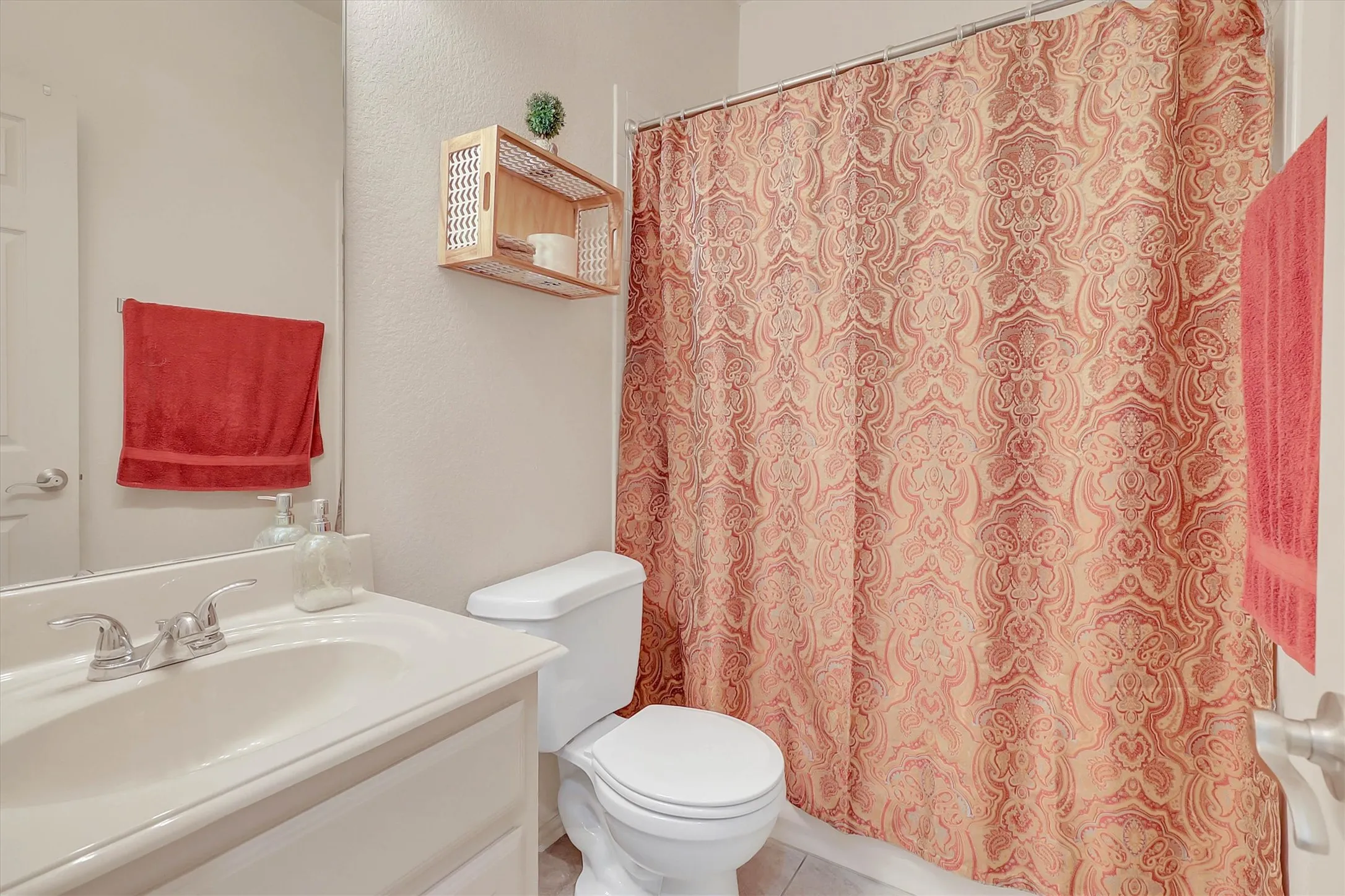 Bathroom featuring a single vanity with chrome fixtures, a toilet, a shower/tub combination with a patterned shower curtain, and a wall-mounted shelving unit