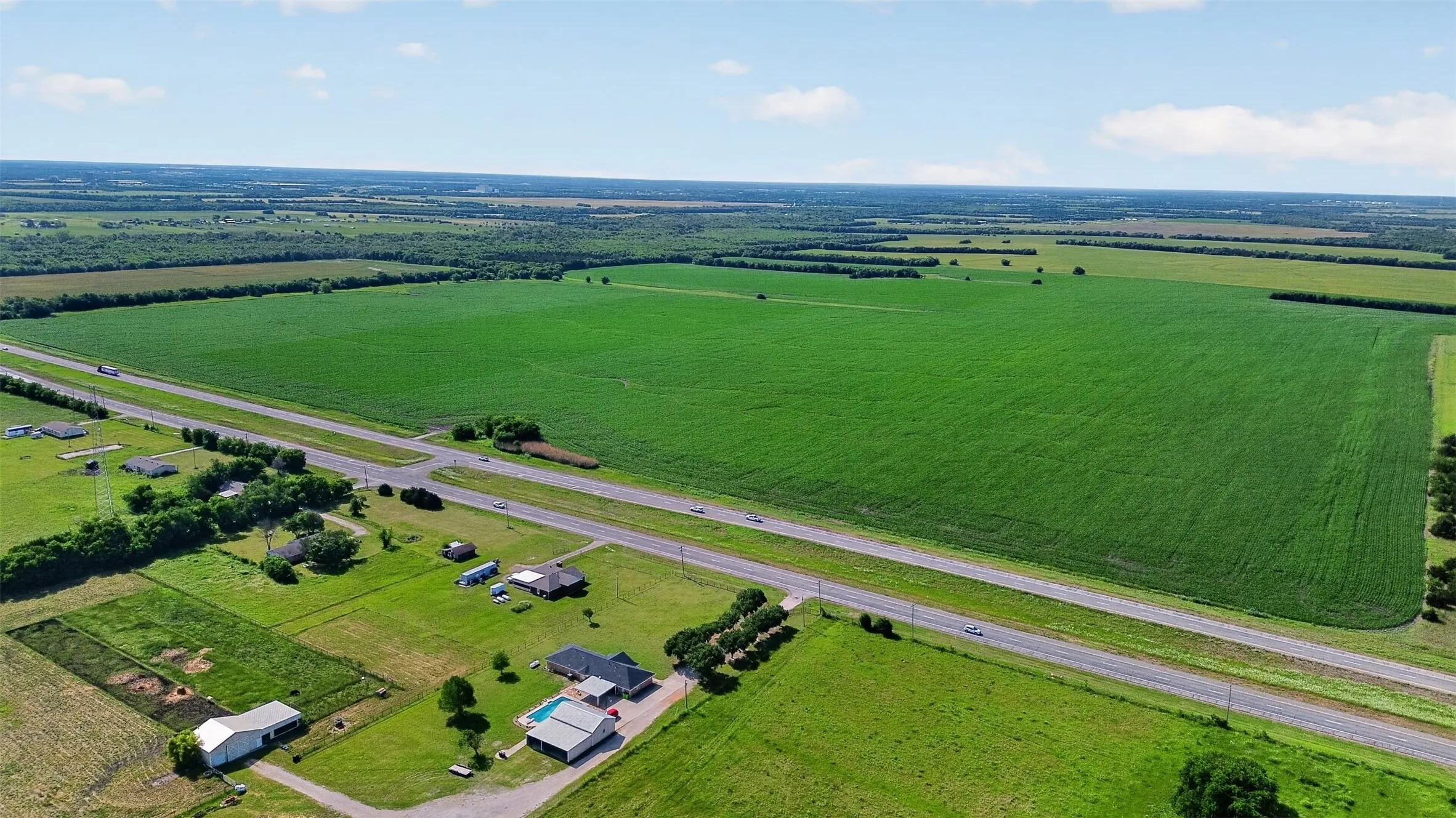 Expansive agricultural land featuring a large green field, a paved road traversing the property, and scattered trees