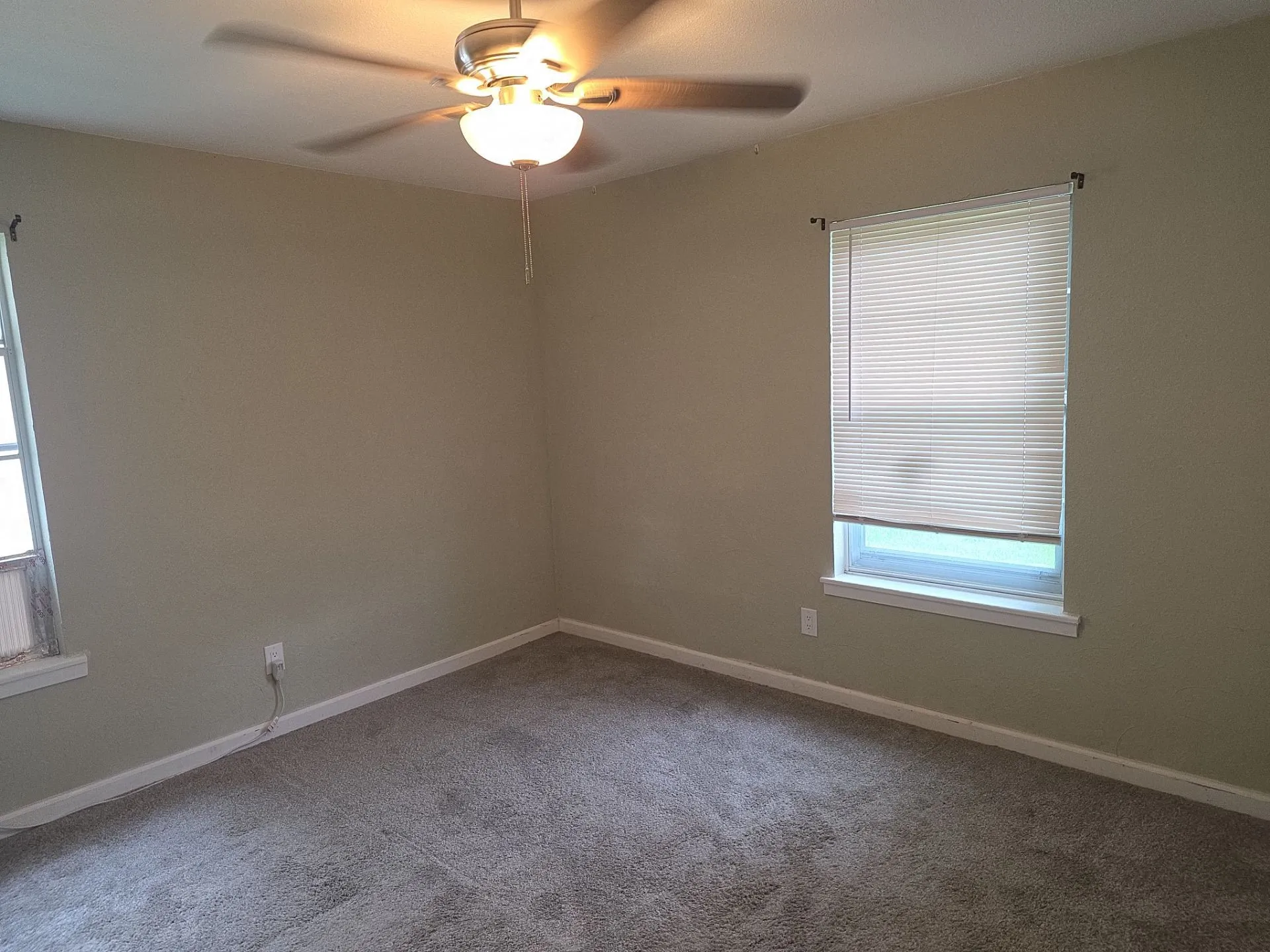 Carpeted room featuring a ceiling fan with light fixture, two windows with blinds, and neutral wall tones
