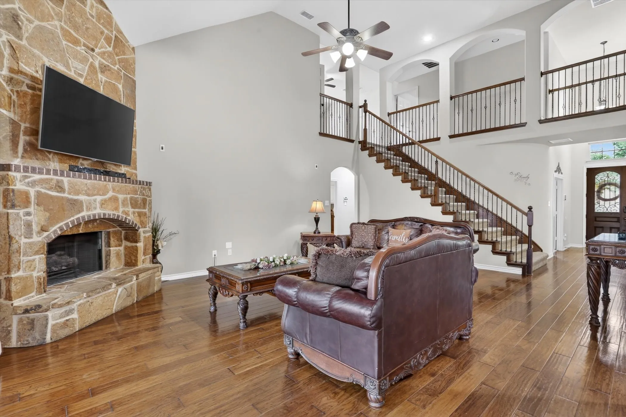 Two-story living area featuring a stone fireplace, wood-finish flooring, and a grand staircase with dark wood railings