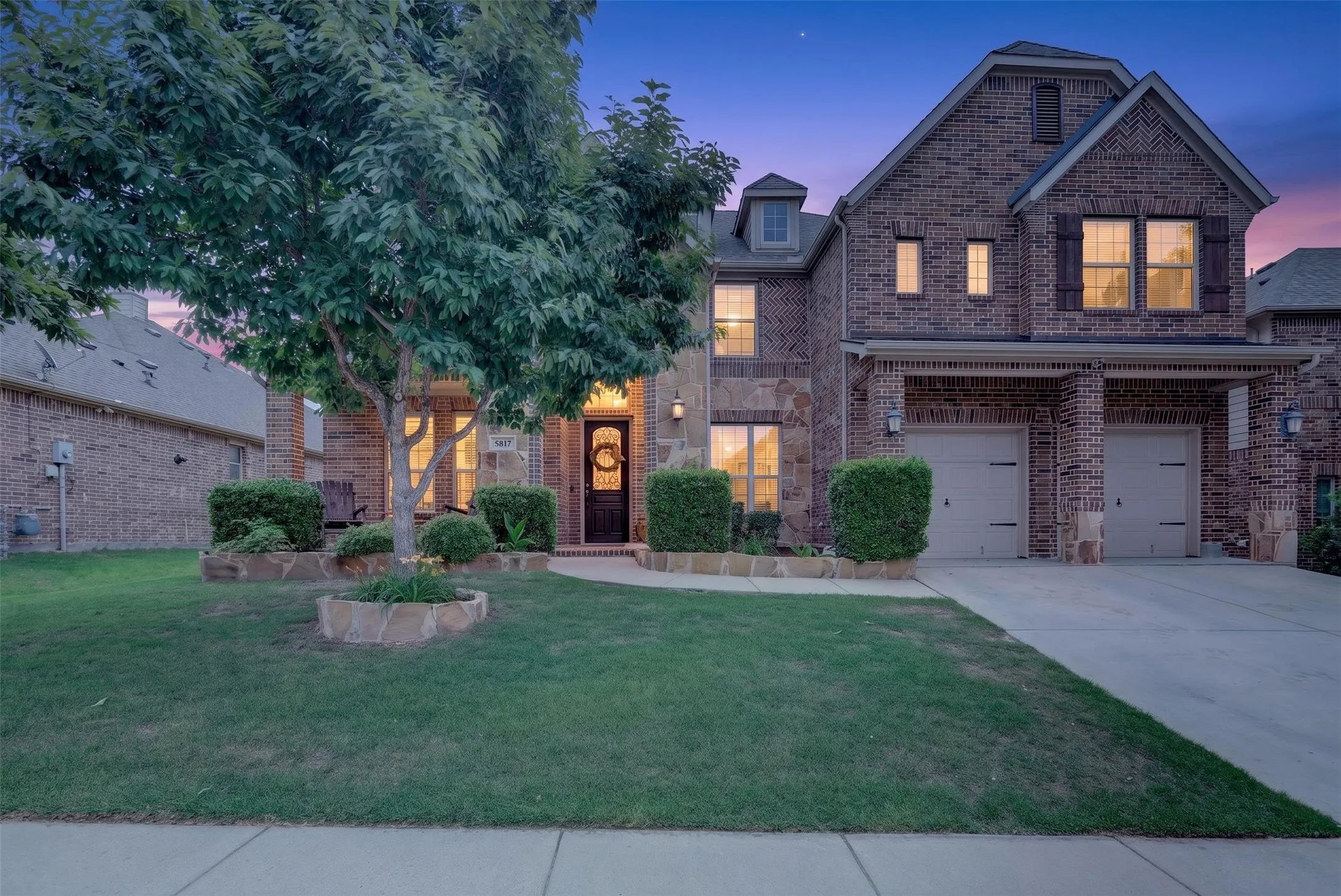 Brick and stone facade, two-car garage, mature front yard tree, stone-edged planter beds, and a covered front entry