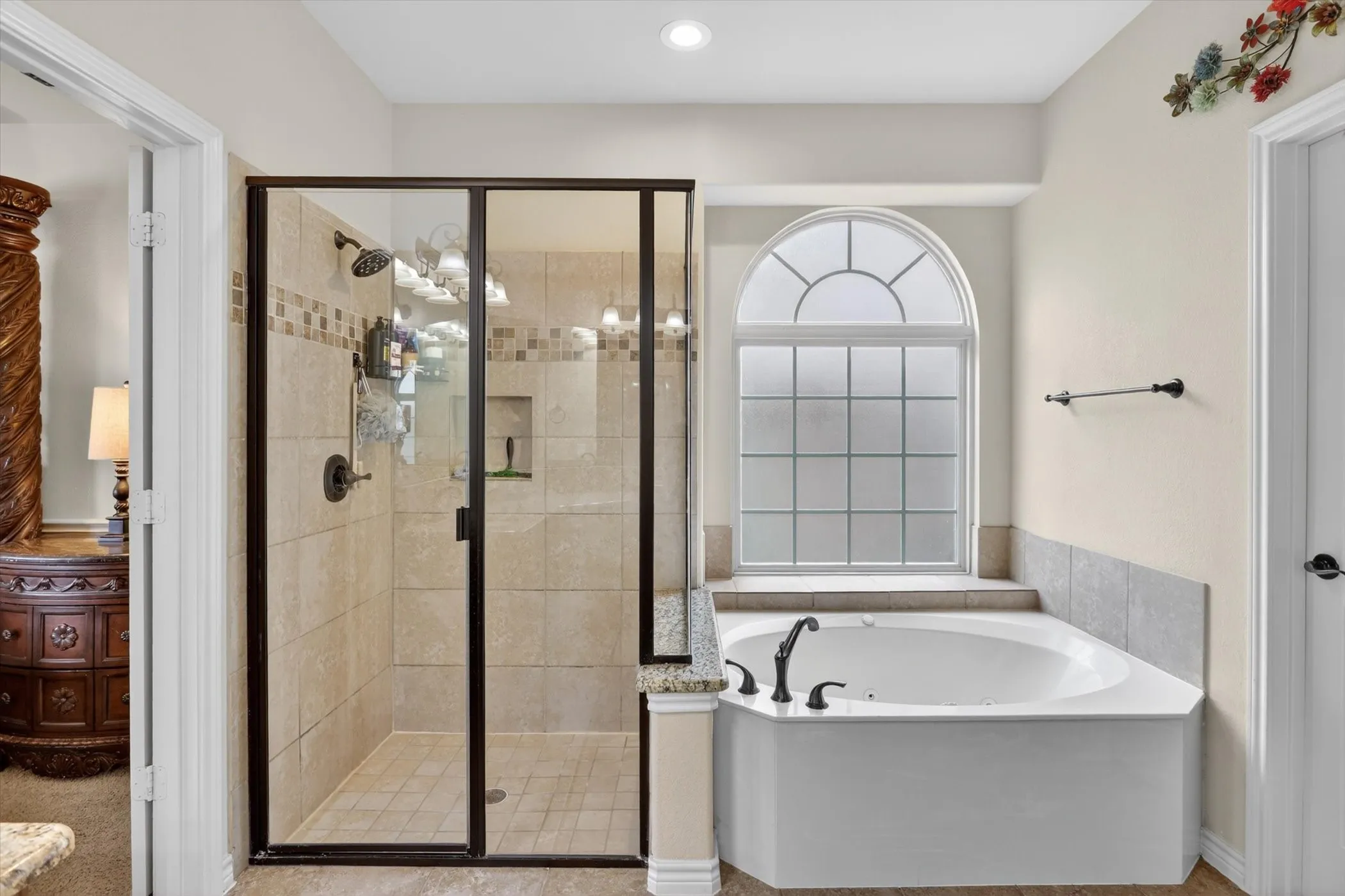 Spacious bathroom featuring a glass-enclosed shower with tile surround and an integrated niche, a corner soaking tub, and a decorative arched window with frosted panes