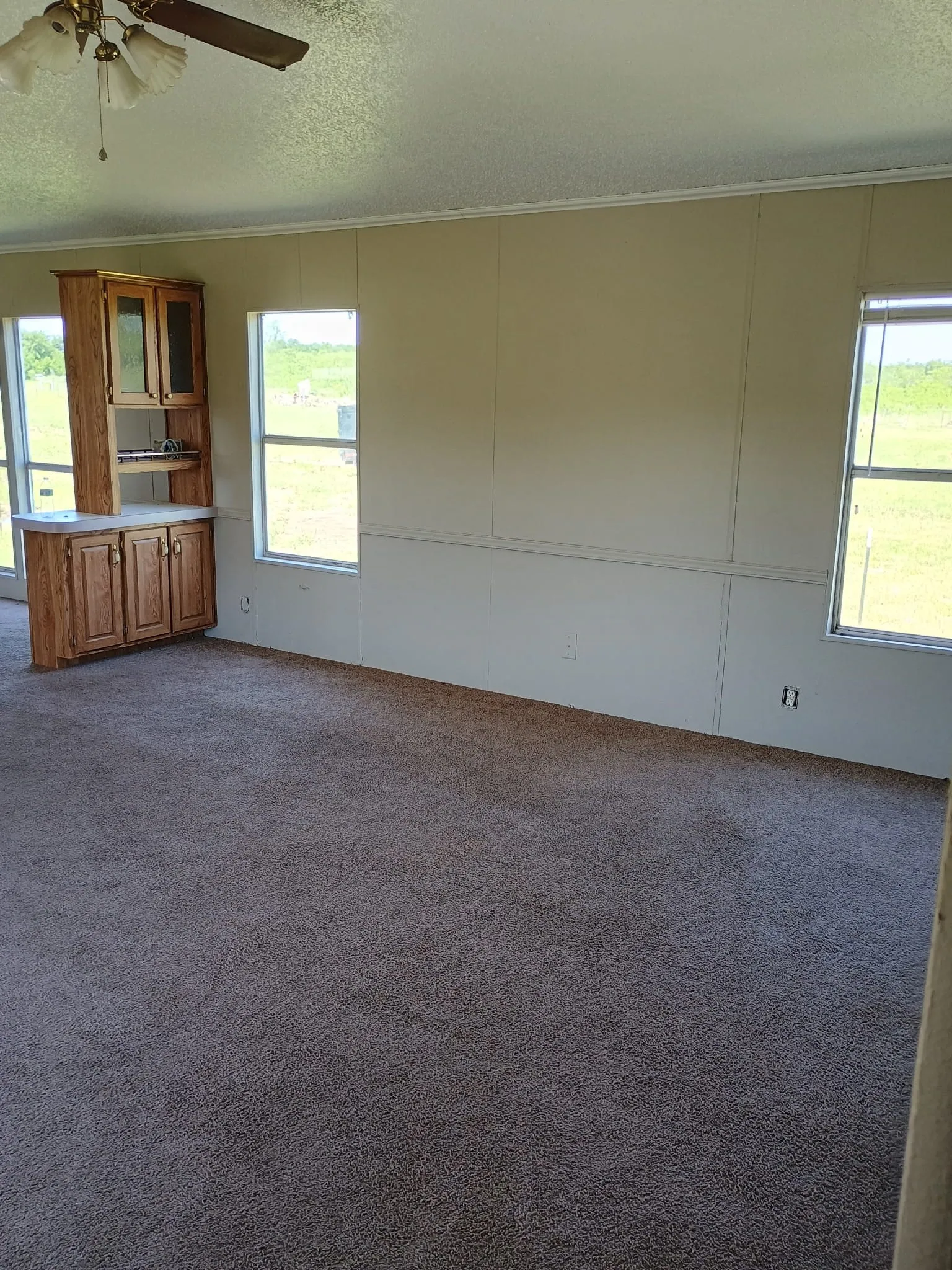 Spacious room featuring neutral wall tones, multiple windows providing natural illumination, and a built-in wood-finish cabinet unit with glass-front upper doors