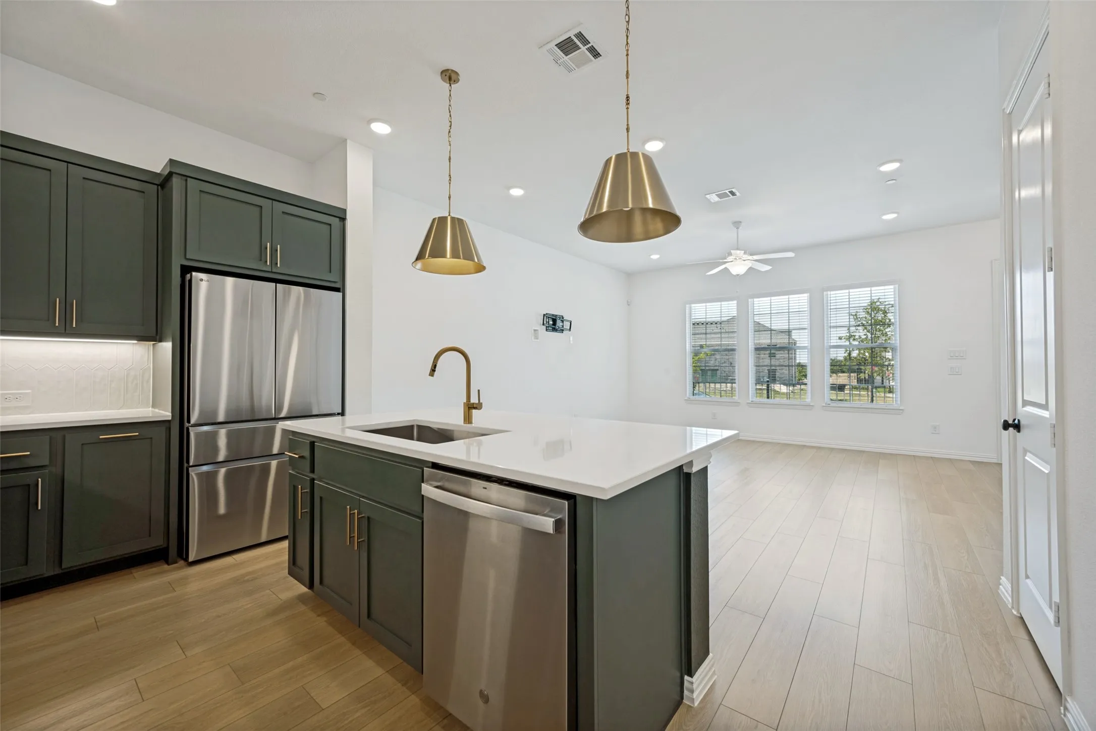 Open-concept kitchen featuring dark green cabinetry, stainless steel appliances, a center island with a sink and brass faucet, and overhead pendant lighting