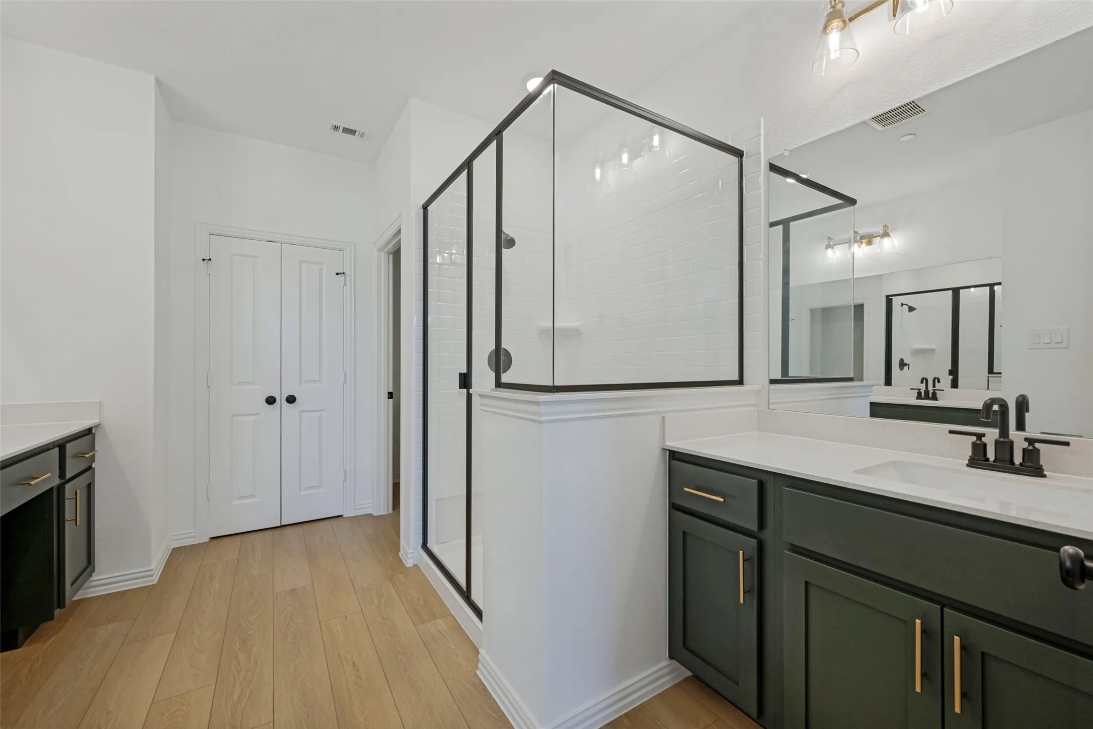 Primary Bathroom featuring a custom walk-in shower enclosure with black metal framing and glass panels, dual vanity with dark cabinetry and brushed gold hardware, quartz countertop, and wood-finish flooring throughout