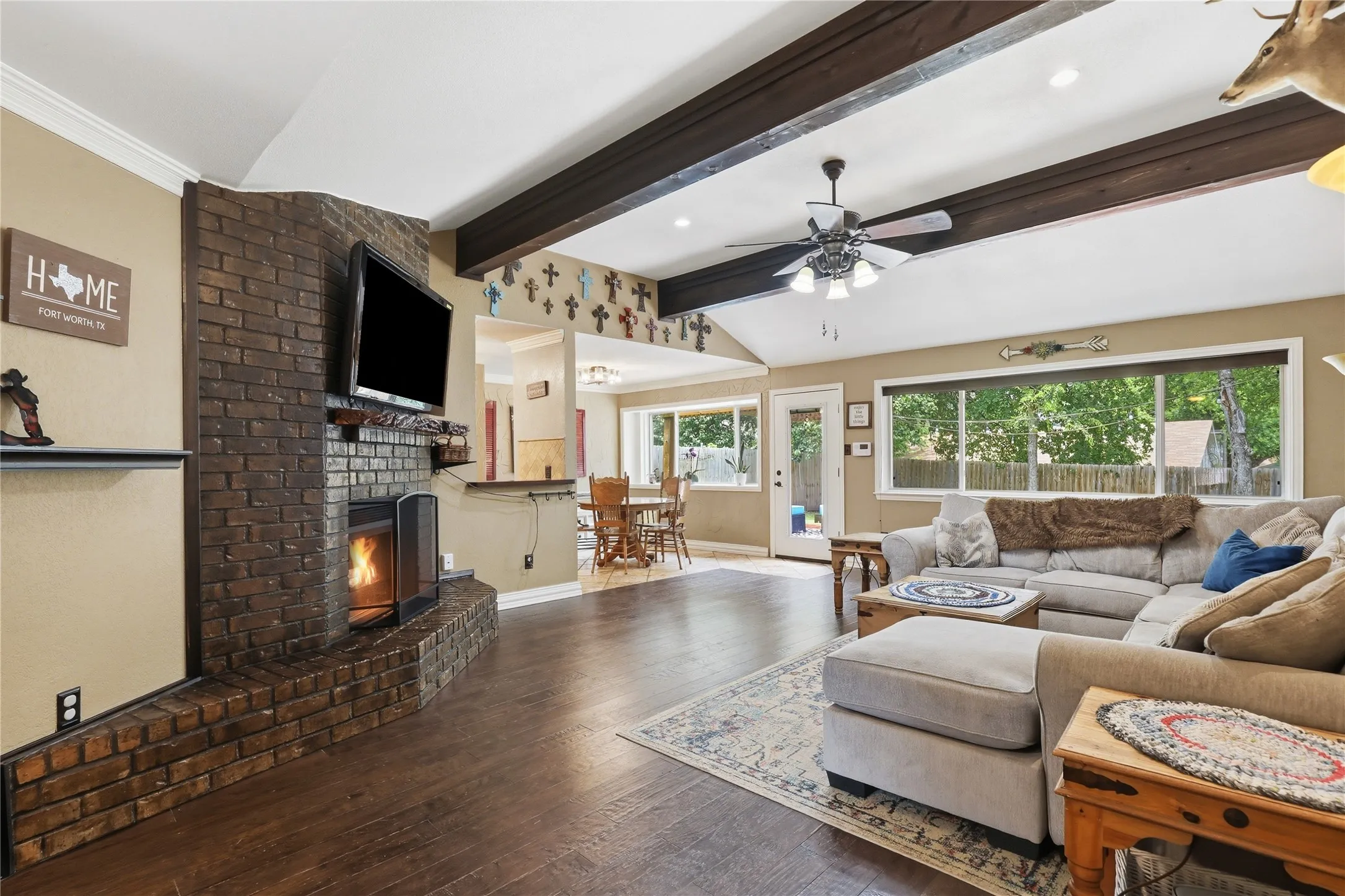 Spacious living area featuring a prominent brick fireplace with a built-in firebox, rich wood-finish flooring, exposed ceiling beams, and expansive windows offering abundant natural light