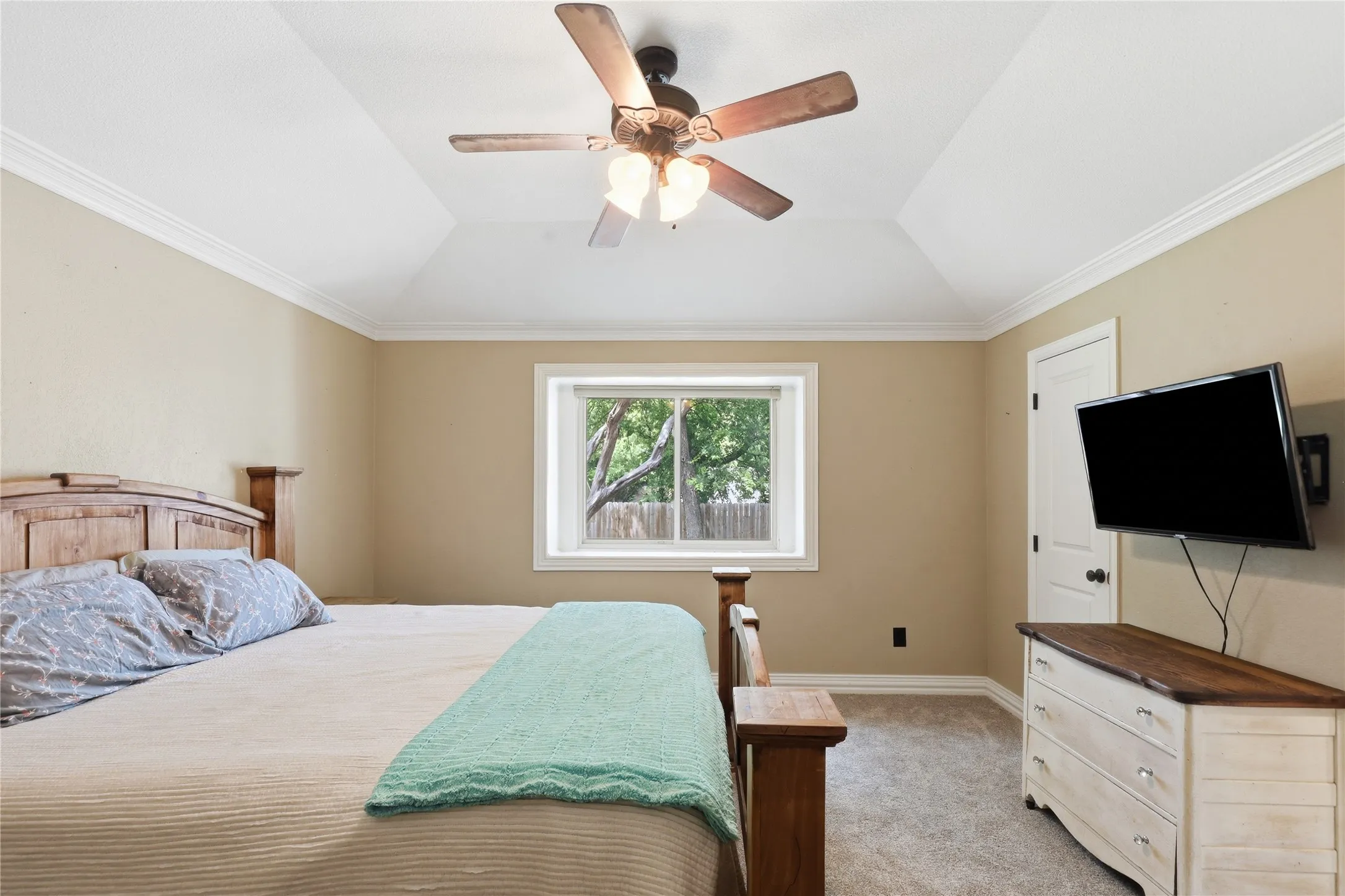 Tray ceiling with crown molding, ceiling fan with integrated lighting, framed window, light-colored carpeting, and a white interior door