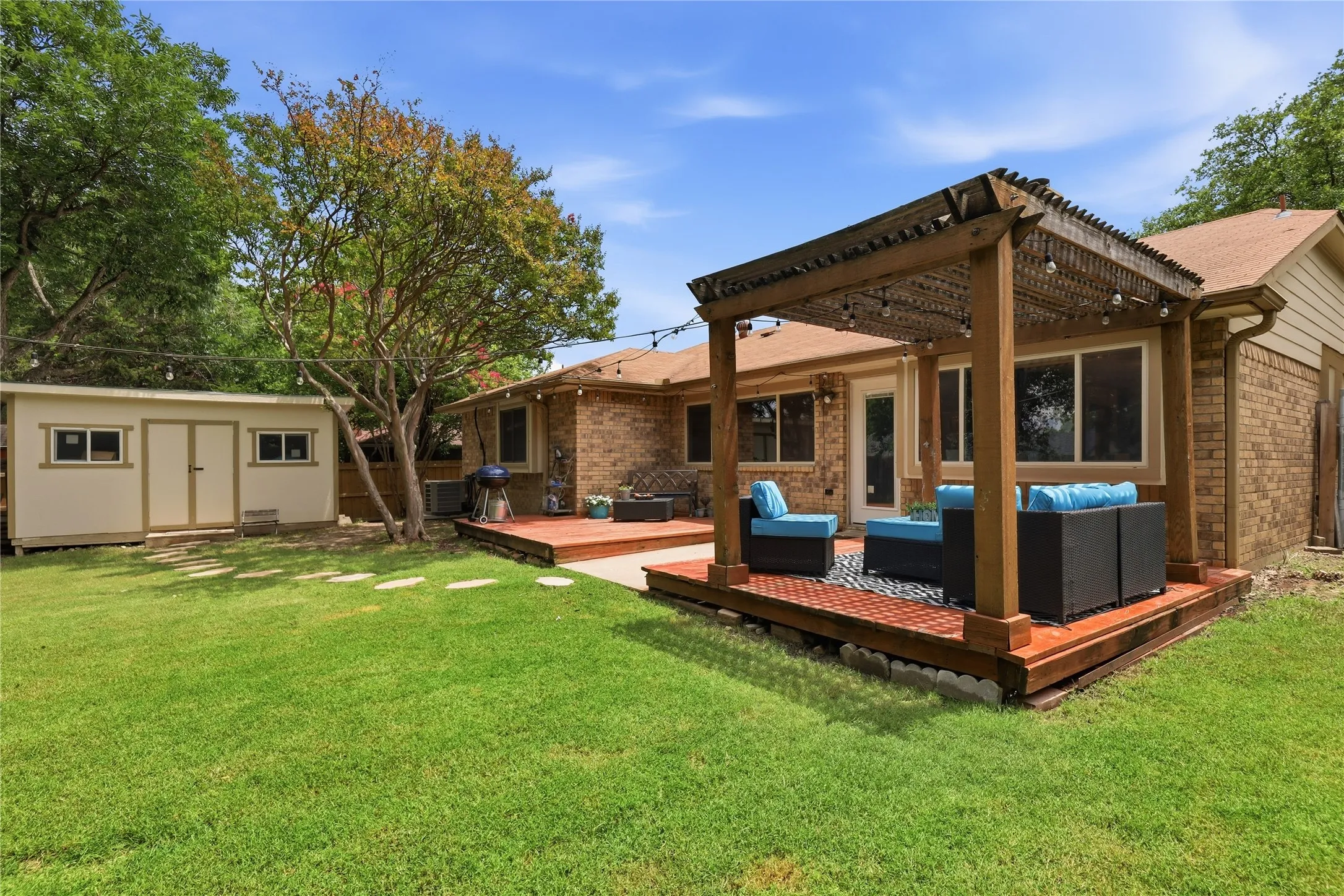 Expansive backyard featuring a large wooden deck with a pergola, a detached storage shed, a manicured lawn, mature trees, and a brick exterior