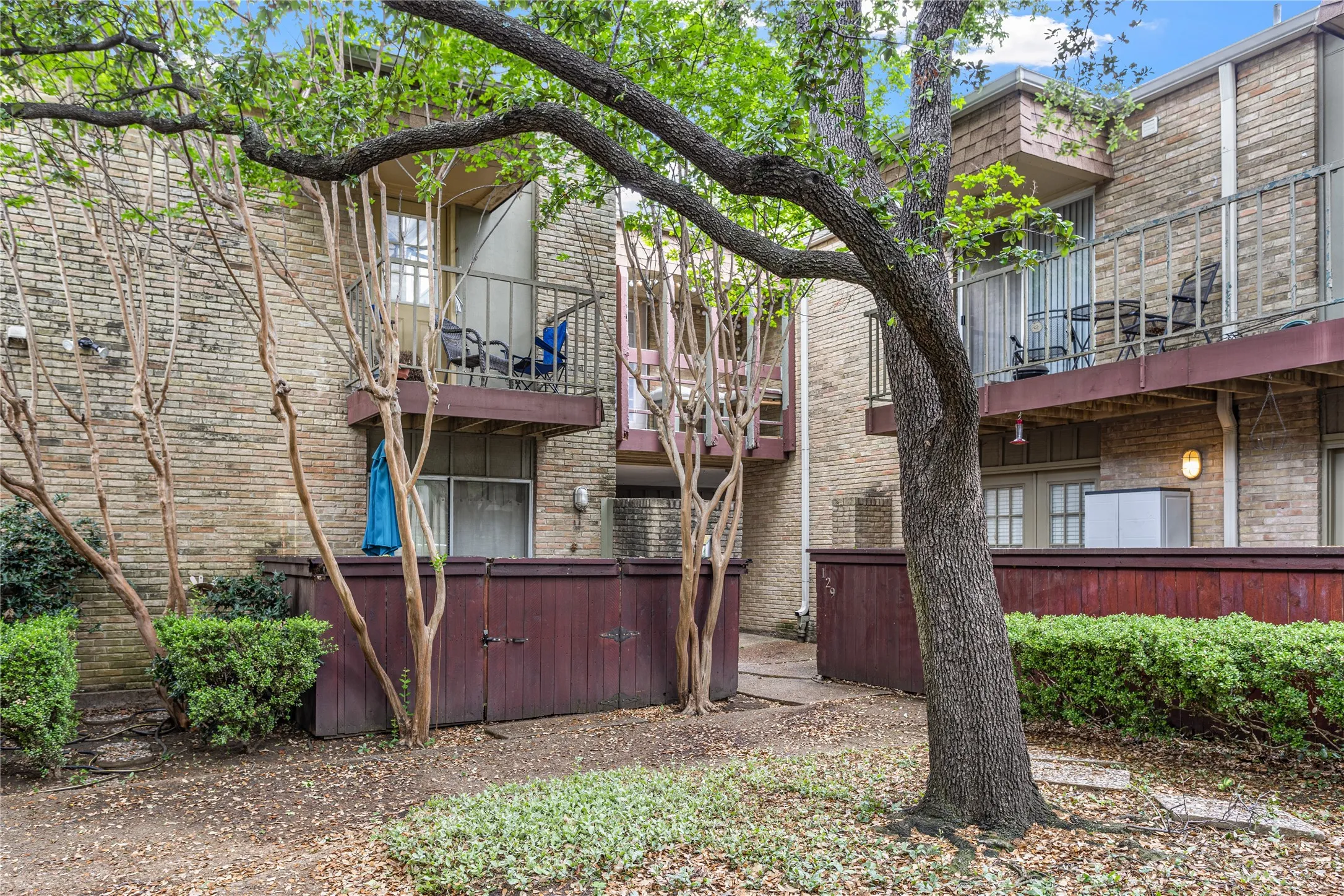 Exterior featuring light-toned brick construction, multiple private balconies with metal railings, mature trees, and established shrubbery