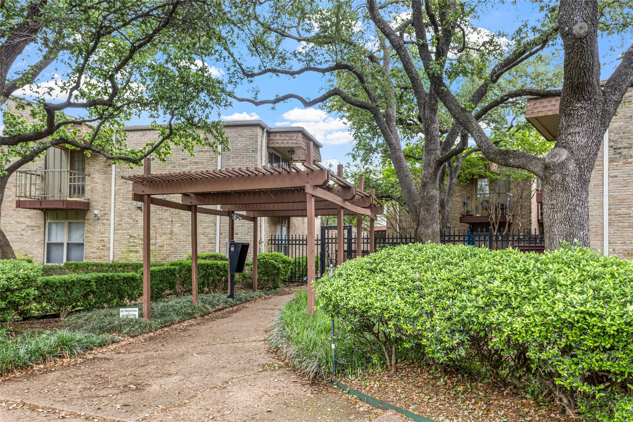 Wood pergola entryway with a paved pathway, surrounded by manicured shrubs and mature trees