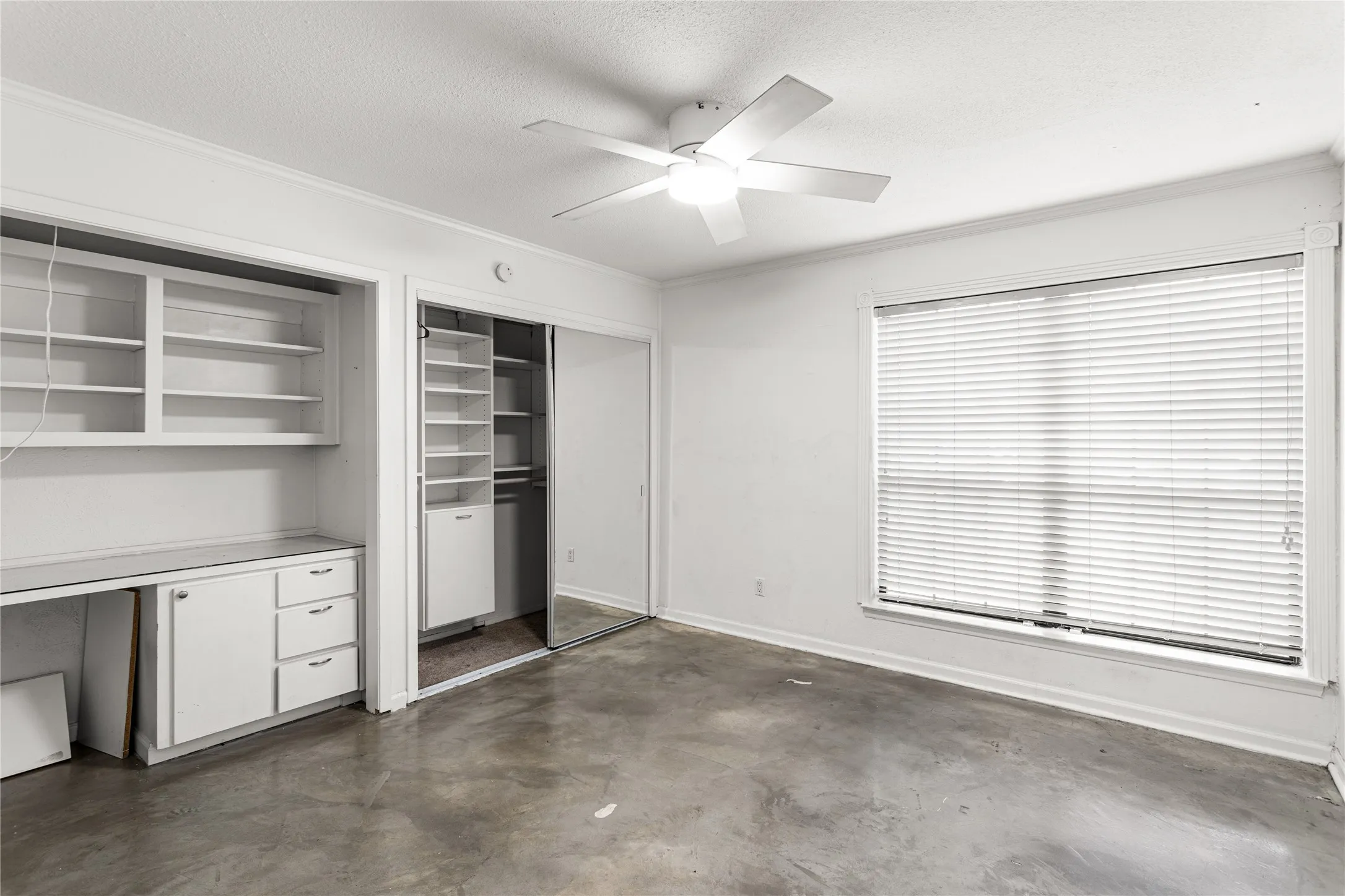 Neutral-toned room featuring a window with blinds, ceiling fan with illumination, and built-in shelving with cabinetry