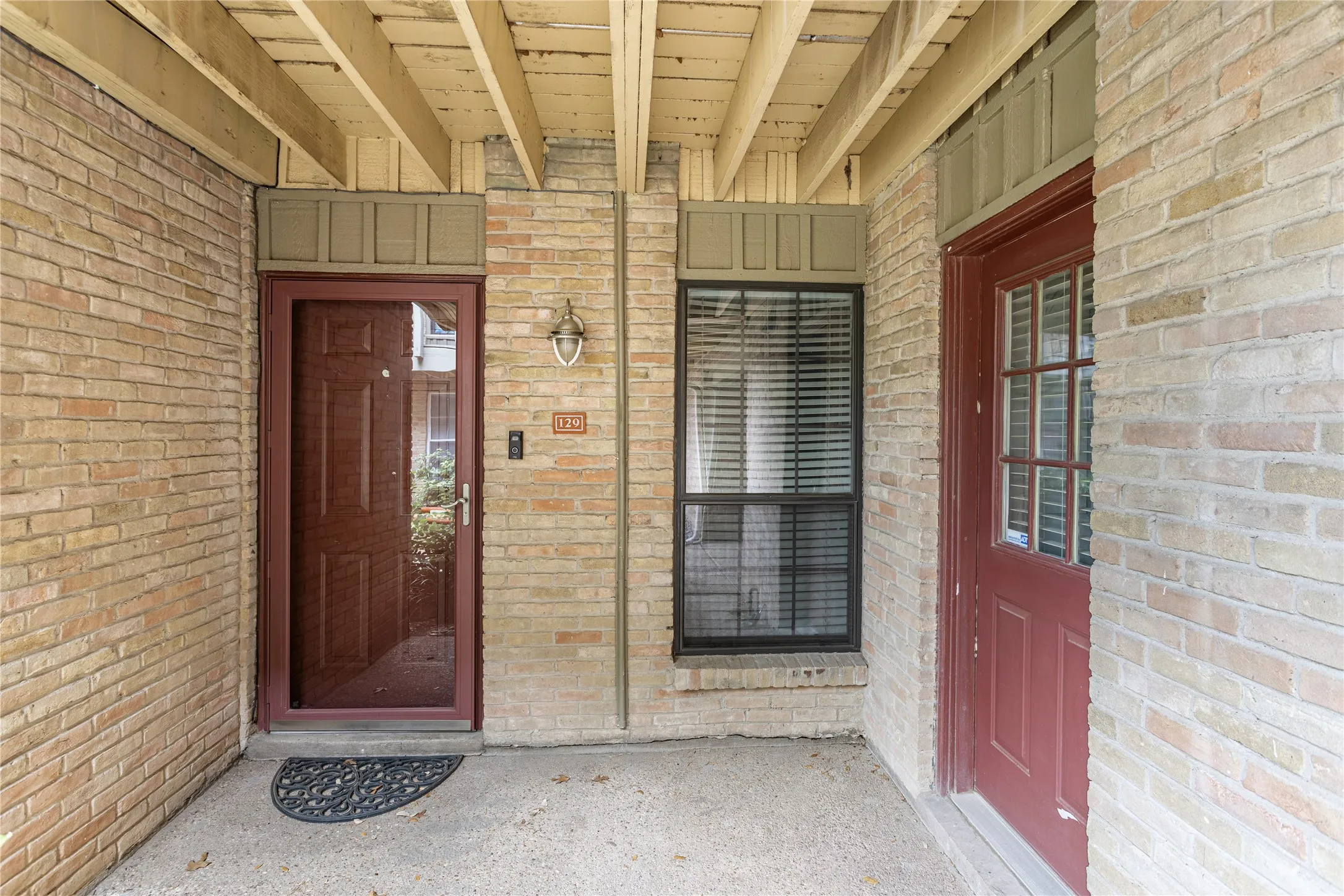 Brick exterior featuring a covered entry, a red-hued main door with a storm door, and a window with horizontal blinds