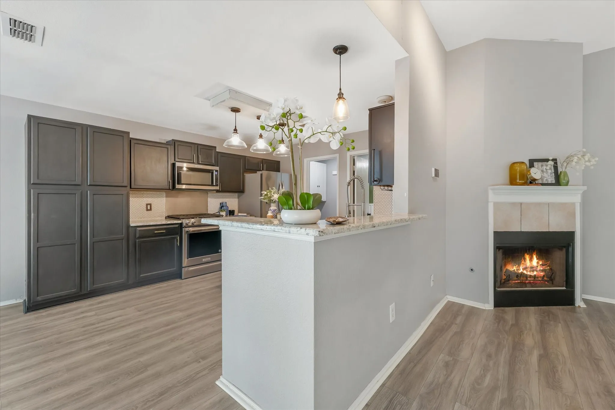 Kitchen and living area featuring wood-finish flooring, dark cabinetry, stainless steel appliances, a breakfast bar with a light-toned countertop, and a fireplace with a tile surround and white mantel