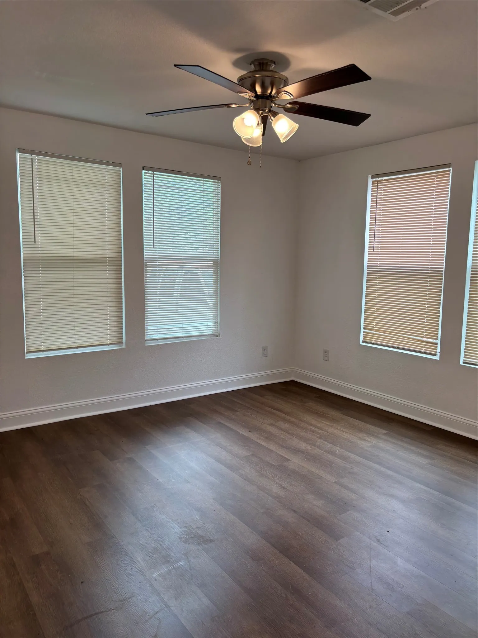 Spacious interior room featuring wood-finish flooring, white walls, and multiple windows with blinds