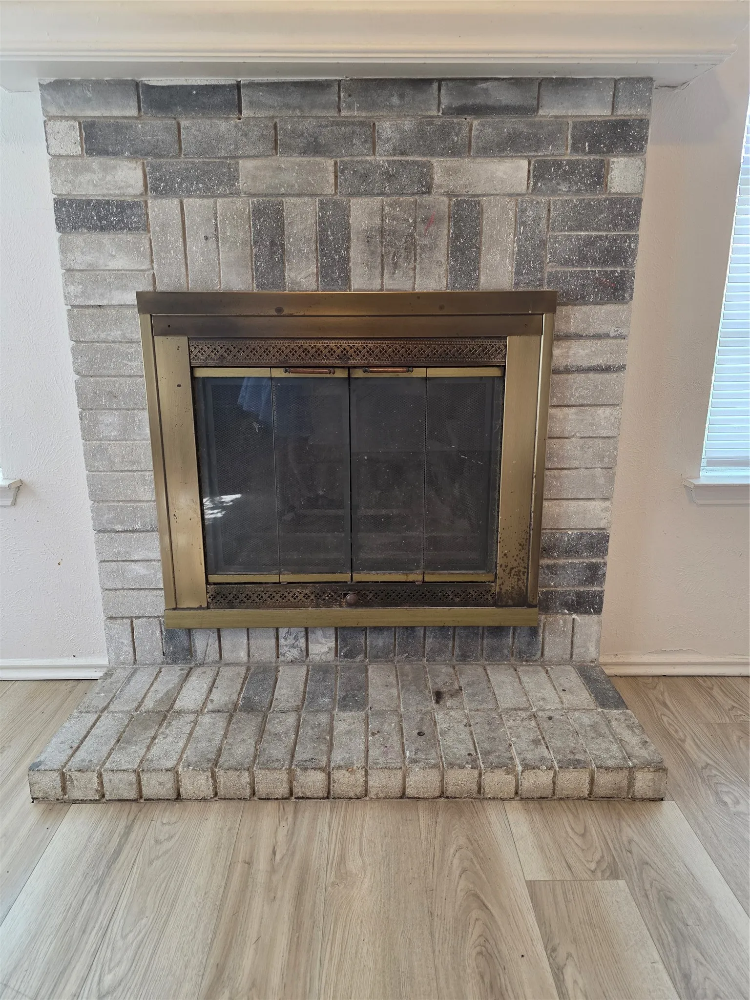 Brick-clad fireplace featuring a brass-toned firebox with glass doors, a brick hearth, and wood-finish flooring
