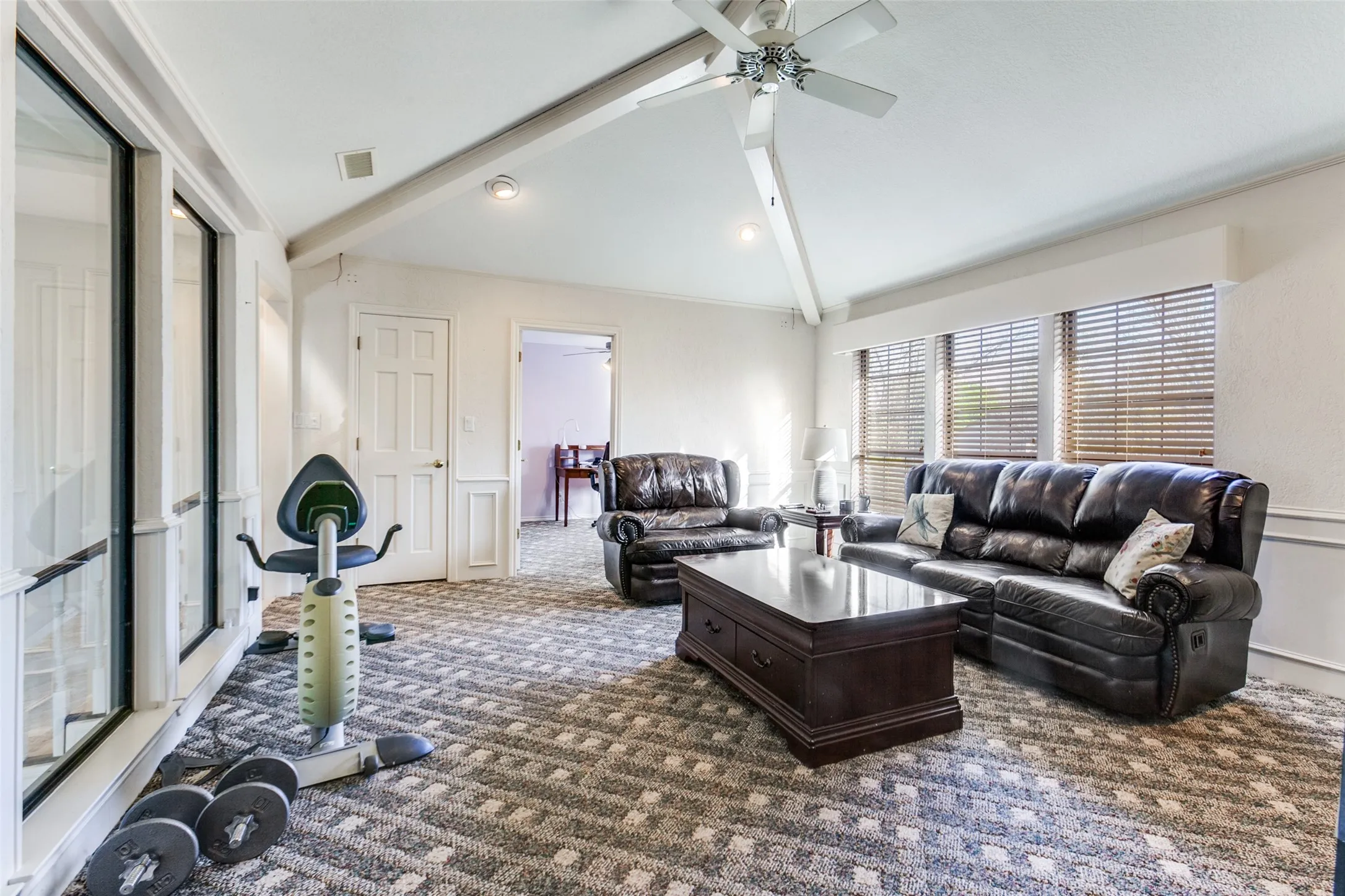 Spacious room featuring vaulted ceilings with exposed beams, large windows with horizontal blinds, patterned carpeting, and recessed lighting