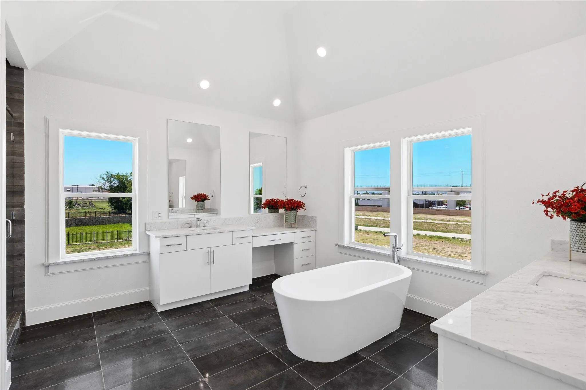 Spacious bathroom featuring a freestanding tub, dual vanities with white cabinetry and light-toned countertops, dark tile flooring, and a vaulted ceiling with recessed lighting