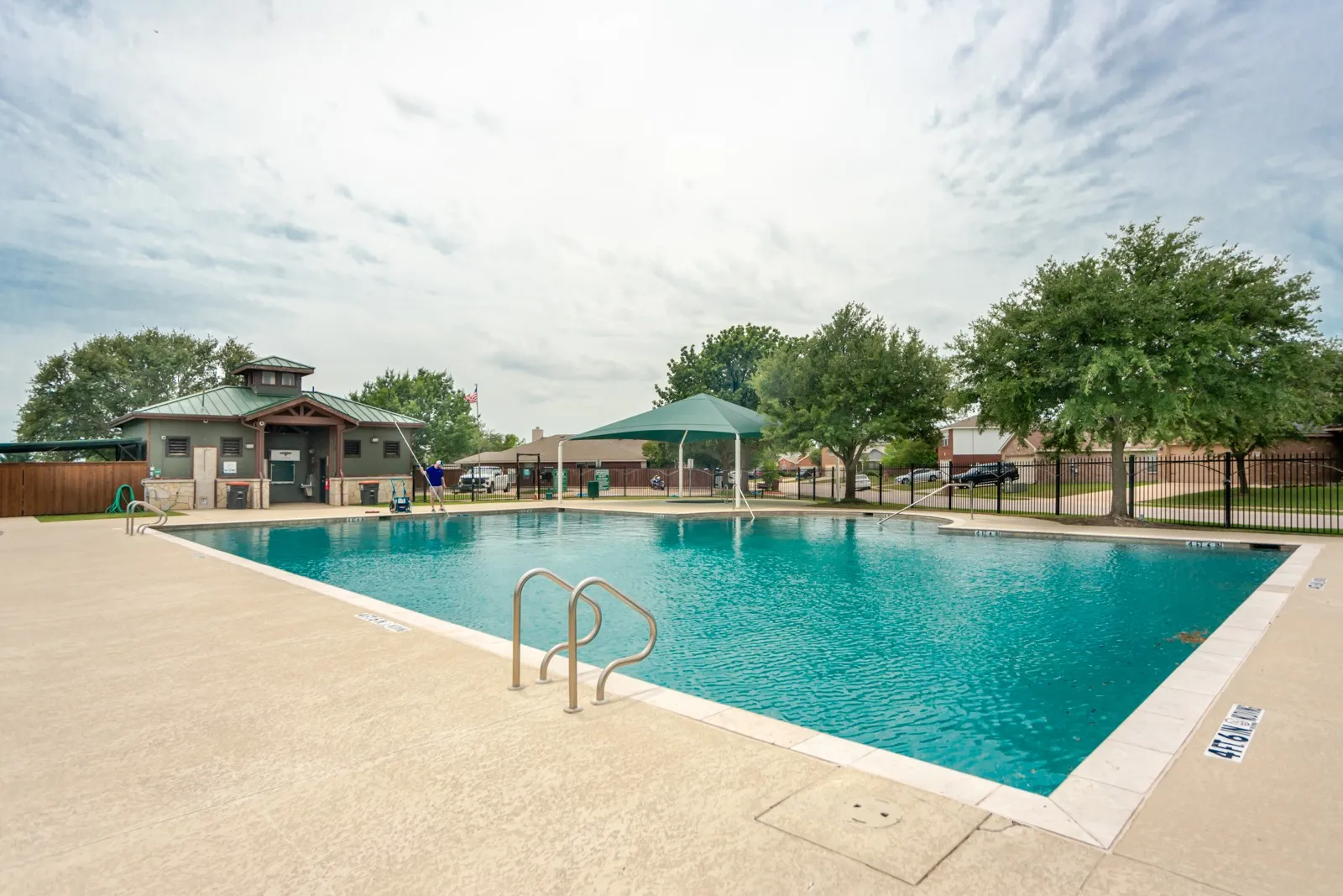 Community swimming pool with concrete decking, perimeter fencing, and a nearby building with a green metal roof