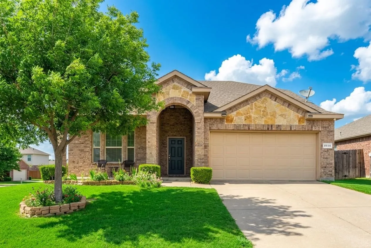 Brick and stone exterior featuring an arched entry, a two-car garage, a mature front tree with a brick-lined flower bed, and a concrete driveway