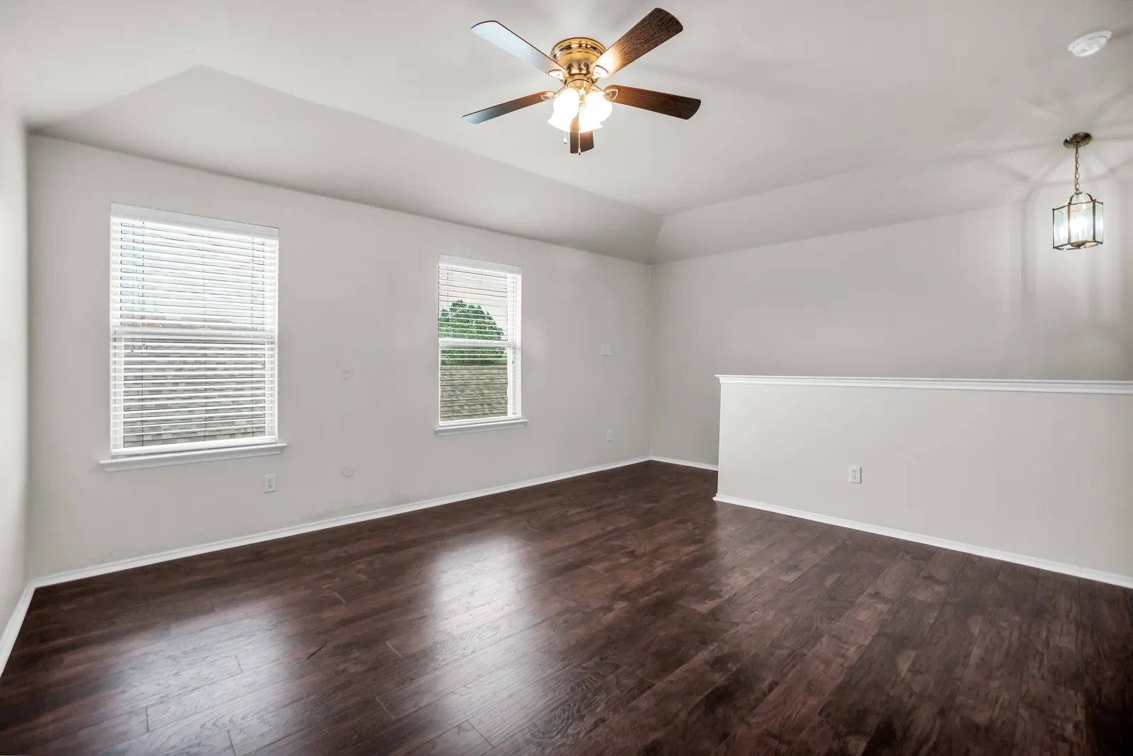Spacious room featuring rich wood-finish flooring, a ceiling fan, and two windows with blinds