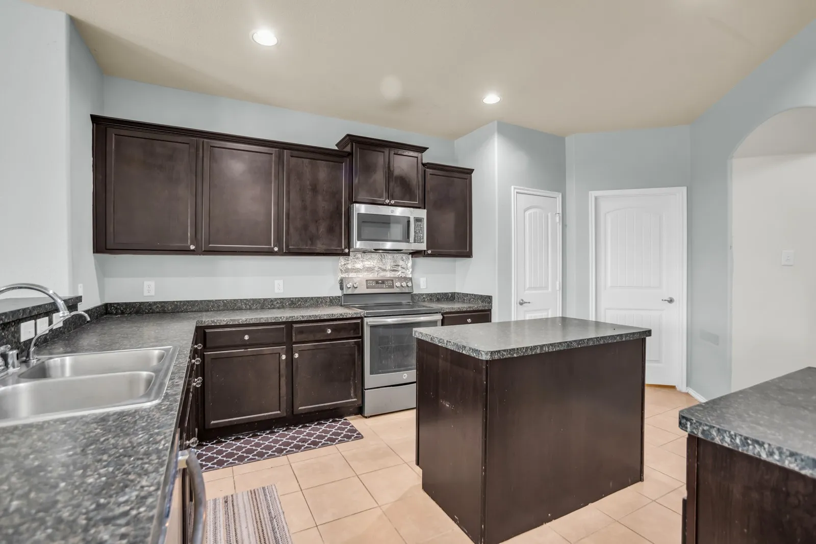 Kitchen featuring dark wood cabinetry, stainless steel appliances, a double basin sink, an island with a stone-finish countertop, and tile flooring