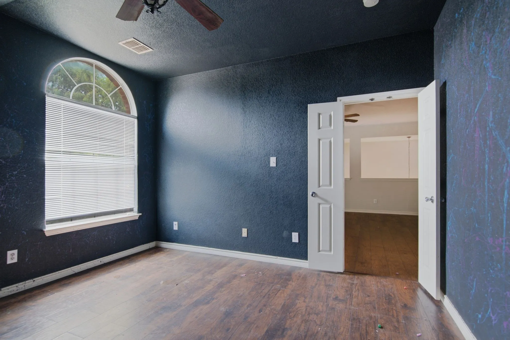 Room featuring dark textured walls, wood-finish flooring, a white arched window with blinds, and a ceiling fan with a light fixture