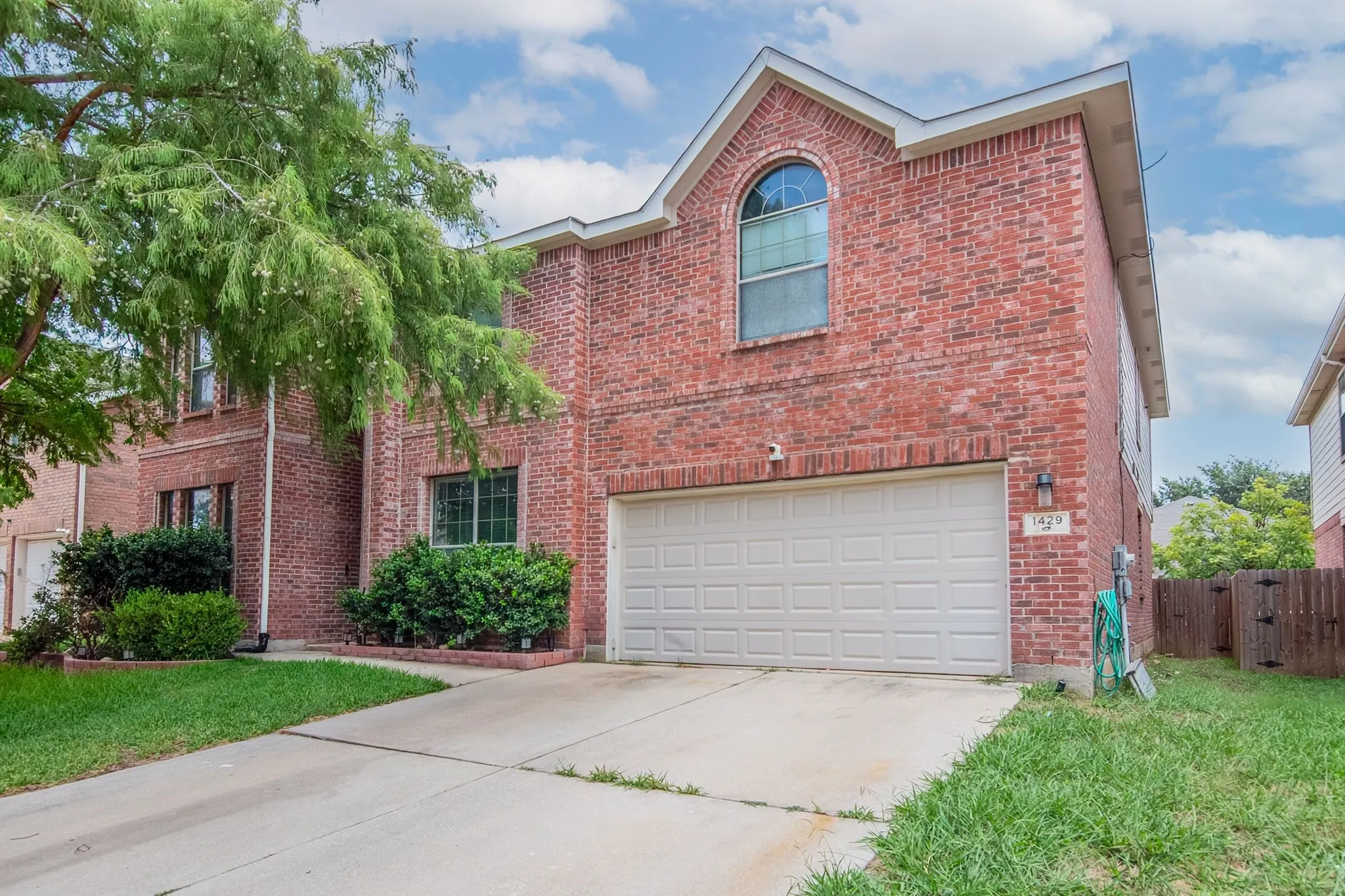 Brick facade residence with an integrated garage, arched window, mature tree, and concrete driveway