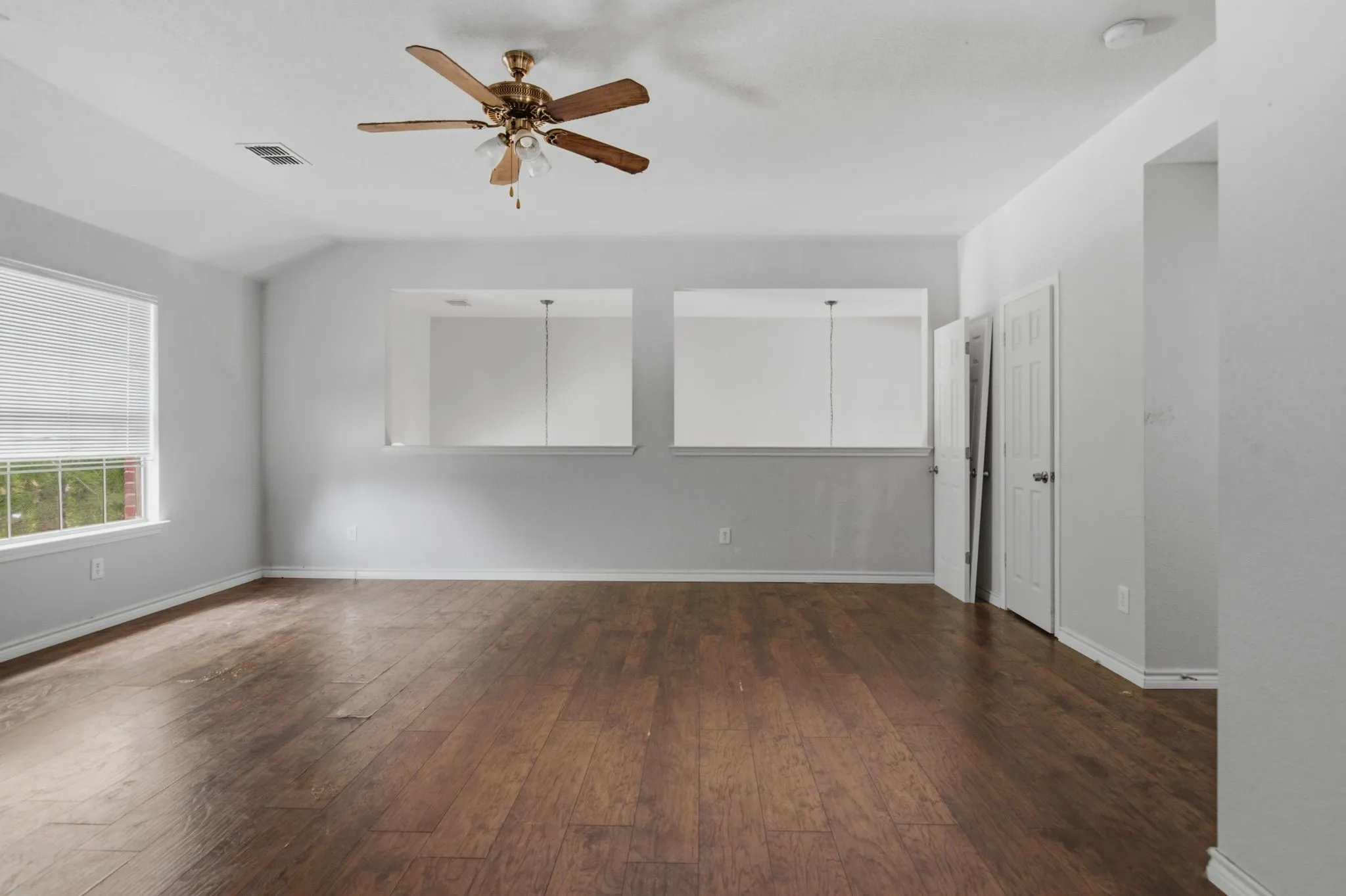 Spacious room featuring wood-finish flooring, two large windows with blinds, and a brass-tone ceiling fan with integrated lighting