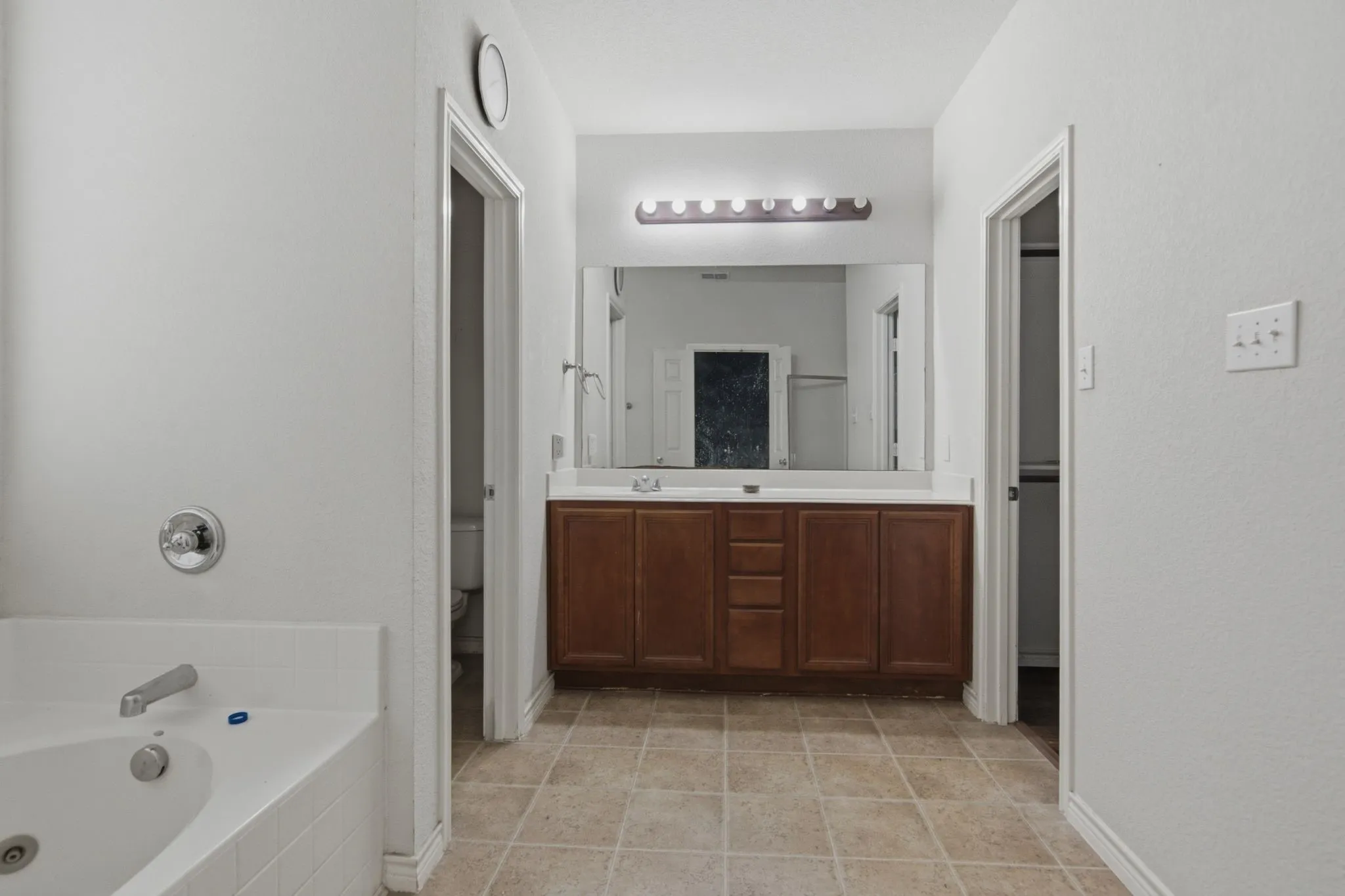 Bathroom featuring a built-in tub, a wood-finish vanity with a double sink, a large mirror, and tile flooring