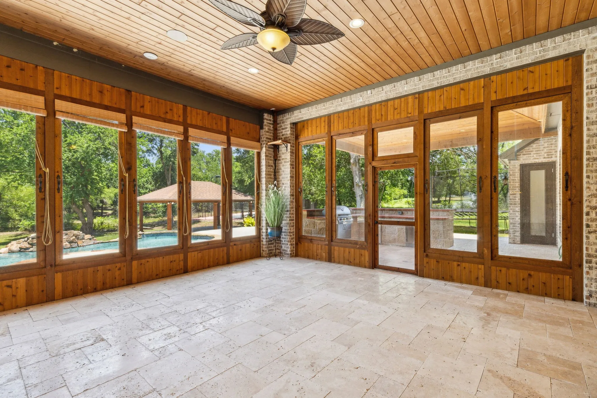 Enclosed patio featuring wood-paneled walls with large glass inserts, light-toned stone tile flooring, and a wood-paneled ceiling with a ceiling fan